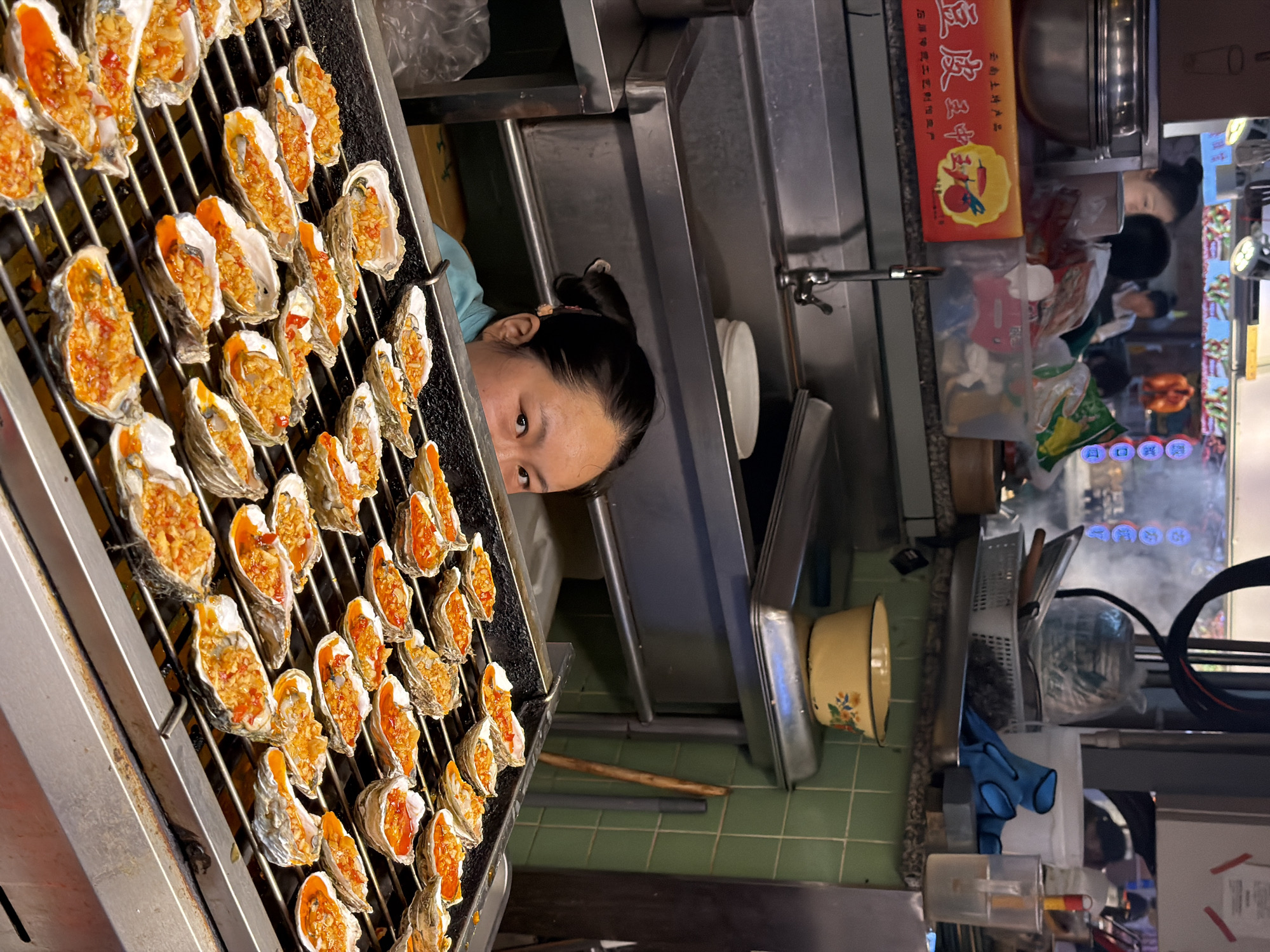 A close-up, low-angle shot captures a woman looking over a grill filled with many oysters topped with a mixture of minced garlic and chili. The oysters are arranged in rows on a metal grate, with some appearing to be actively cooking. The woman, wearing a light blue top, has dark hair pulled back and is peeking over the grill, her eyes visible. The background is a kitchen or food stall environment, with stainless steel surfaces, sinks, and various containers. A red box with Chinese characters is visible on a shelf above the sink. The scene is likely a food vendor in Yuzhong District, China, given the context provided.