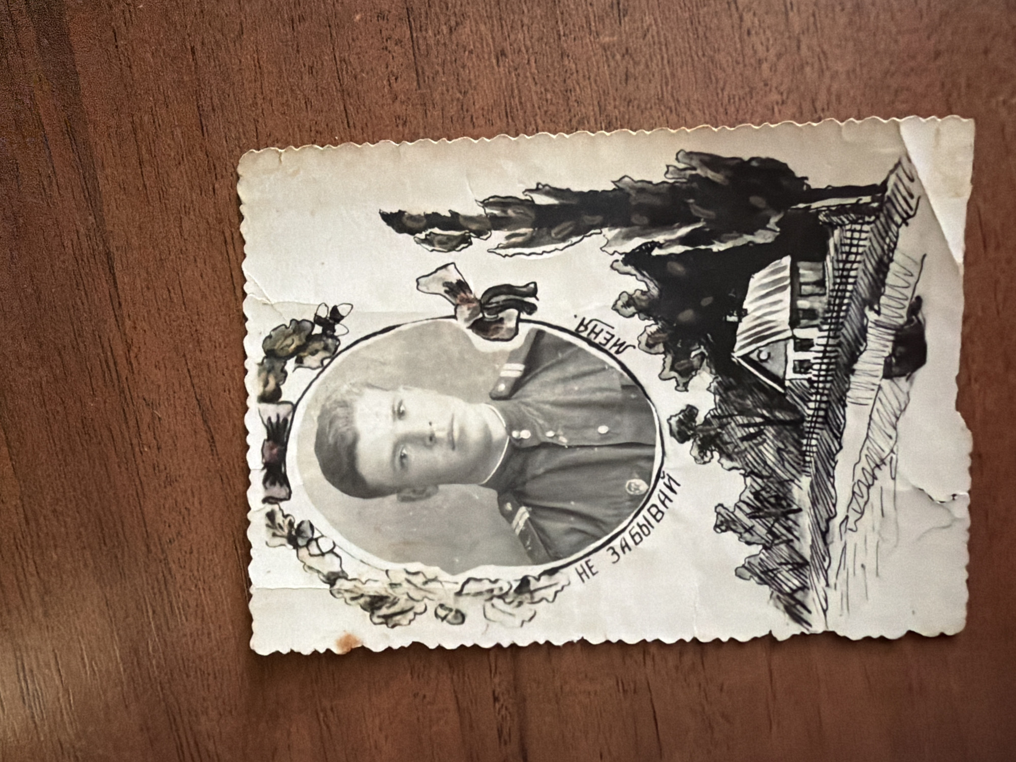 A black and white portrait photo is placed on a wooden surface. The photo depicts a young man in a military uniform. He has short, dark hair and is looking directly at the camera with a neutral expression. He is wearing a dark uniform with a high collar and what appears to be insignia on the shoulders. The portrait is framed by a decorative oval border, with floral motifs and a bow on the left side. Below the portrait, there is a handwritten inscription in Russian, which appears to say "НЕ ЗАБЫВАЙ МЕНЯ" (Don't forget me). The bottom portion of the photo features a sketch of a rural landscape, with a house, trees, and a fence. The photo has a decorative, serrated edge along the right side and some minor damage and discoloration, indicating its age.