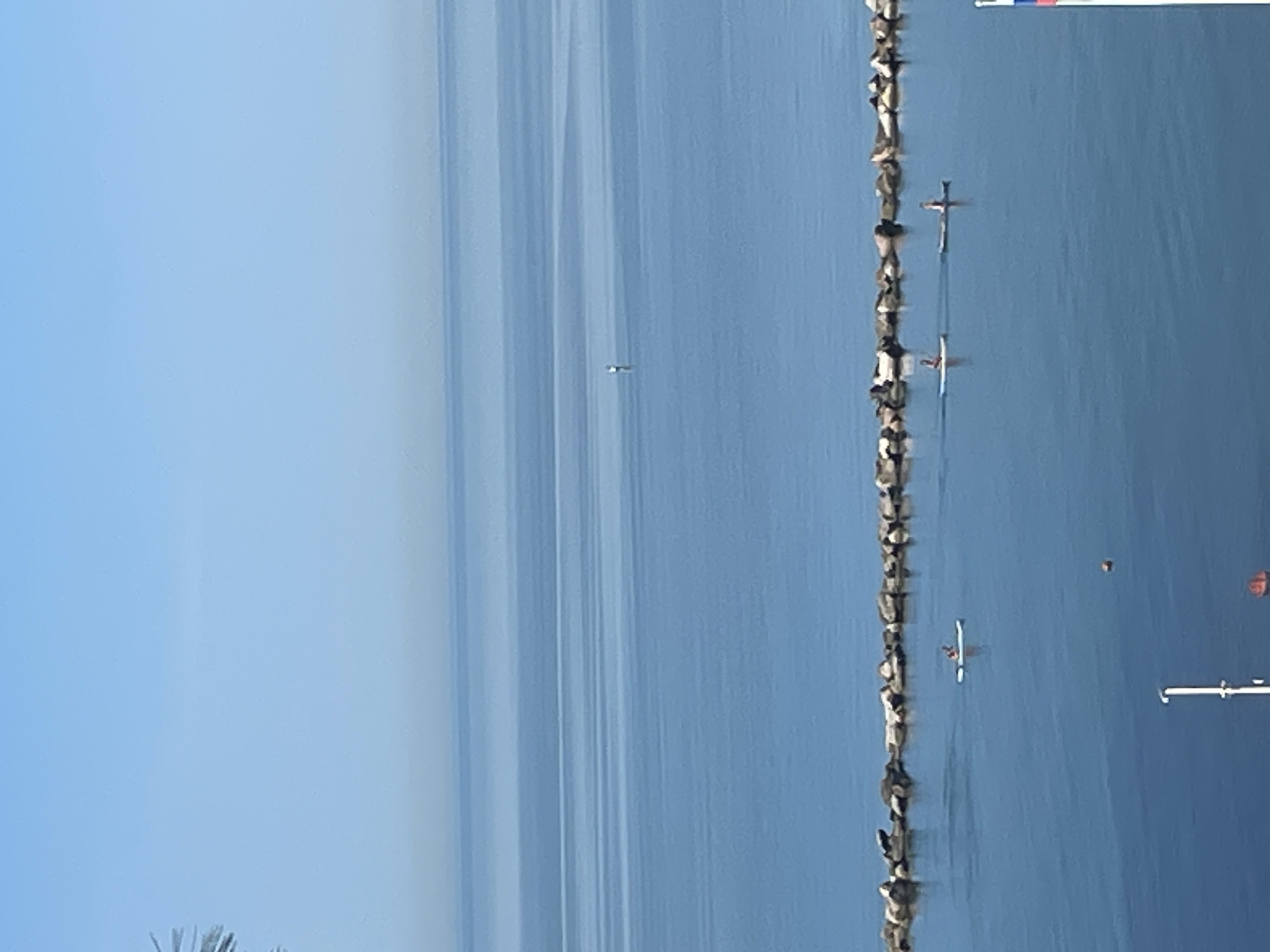 The image depicts a serene coastal landscape, likely captured on a clear day in Sochi, Russia. A vast expanse of calm blue water dominates the scene, meeting a light blue sky at a hazy horizon. The water's surface is relatively smooth, showing subtle variations in color that create faint horizontal bands.
Running horizontally across the middle of the frame is a rugged breakwater or seawall, constructed from numerous rocks, separating the open sea from a presumably shallower area closer to the shore. Several people are engaged in water activities near this structure. At least two individuals are visible on paddleboards or small kayaks; one stands actively paddling near the breakwater, and another is further along it. A third person is seen on a paddleboard or similar craft in the foreground. A very small, distant object in the far water suggests another distant person or boat.
Notable objects include a bright orange buoy floating in the water to the right, and a white pole or structure partially visible in the bottom right corner, hinting at a nearby pier or similar installation. In the top right corner, a small segment of a flag or banner with horizontal red, blue, and white stripes is visible, consistent with the Russian flag, which supports the location context of Sochi.
The scene suggests bright daylight conditions and very calm weather, ideal for recreational watersports. The overall atmosphere is peaceful and leisurely. Blurred foliage can be seen in the bottom left, indicating the presence of land where the photograph was taken.