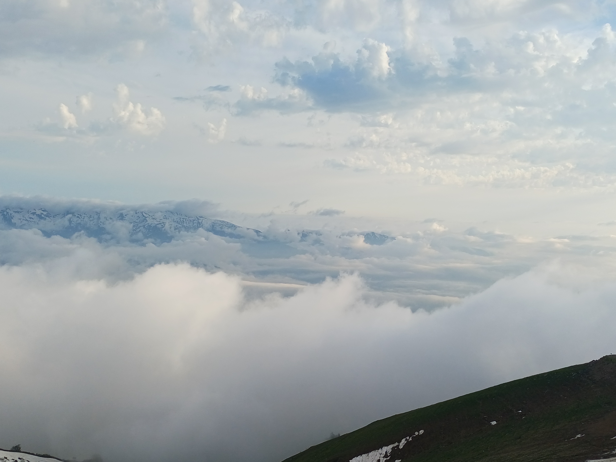 The image captures a panoramic view of a mountainous landscape, likely near Estosadok, Russia, under a cloudy sky. In the foreground, a green hillside slopes upwards from the bottom right, with patches of snow visible on its surface. The midground is dominated by a thick layer of white clouds that obscures the lower parts of the mountains. Above these clouds, the peaks of distant snow-capped mountains are visible, their rugged outlines softened by the atmospheric haze. The sky above is a canvas of varying shades of gray and white, with distinct cloud formations ranging from wispy cirrus to more dense cumulus. The overall impression is one of a serene and majestic natural scene, potentially captured during early morning or late afternoon given the soft lighting.