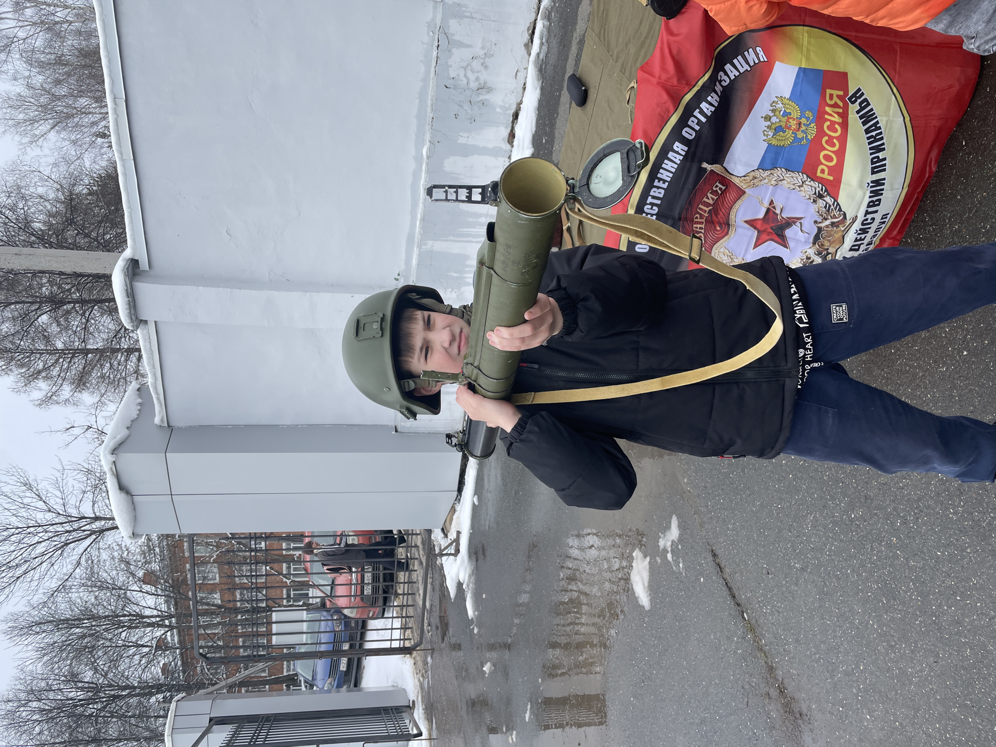 A young boy wearing a military helmet is holding a green-colored tube resembling a rocket launcher. The setting appears to be outdoors near a building, with a small patch of snow on the ground. In the background, a Russian flag with a Soviet star is visible. Cars are parked behind a fence, and the trees are bare, indicating it might be winter or early spring. It is not possible to tell the location is in Sarapul, Russia from the image.