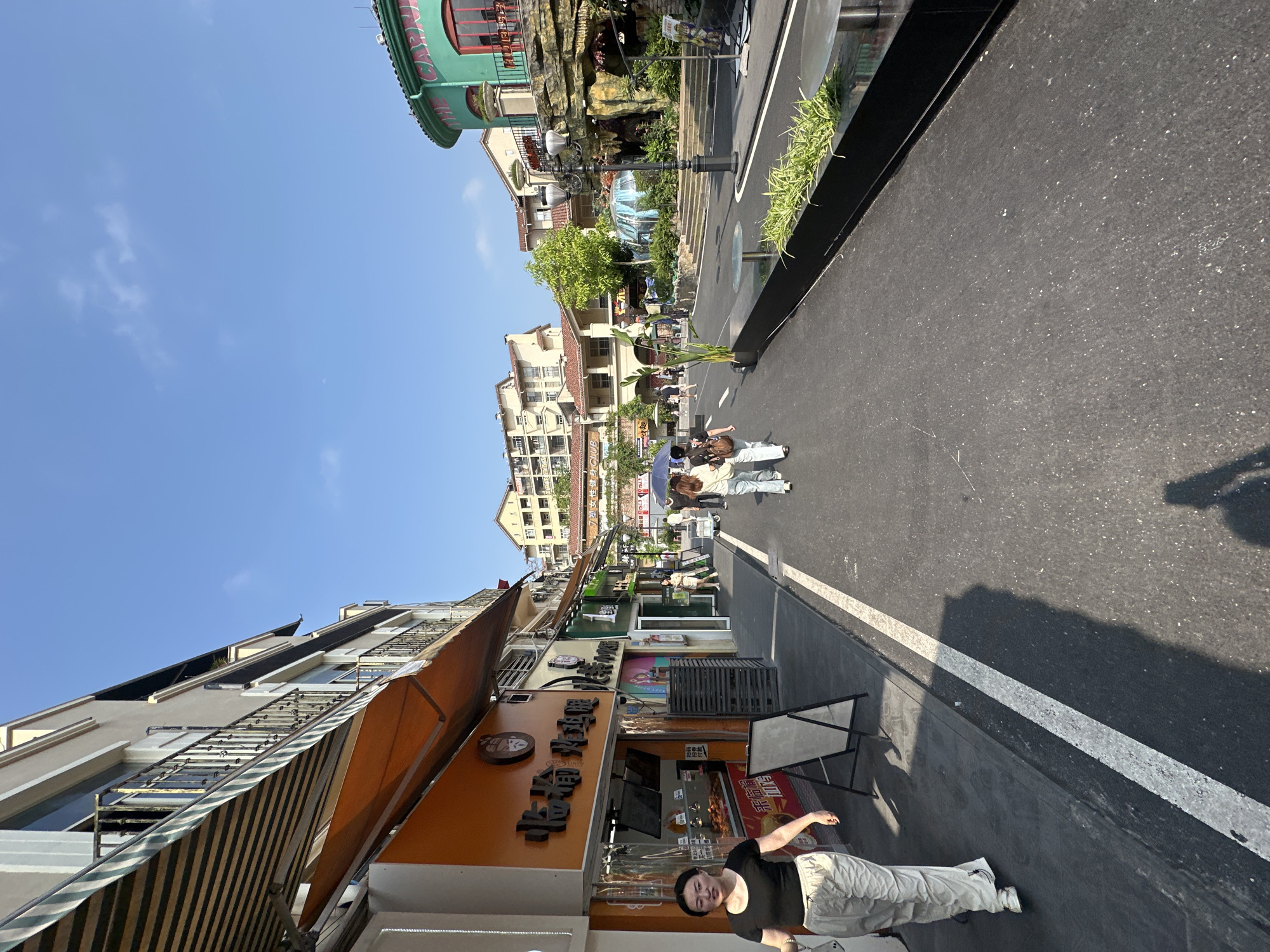 A street view in the Chongqing High-tech Zone, China, reveals buildings along a road. The sky is clear blue. Buildings of various architectural styles line the street. People are walking on the sidewalk and the road. A woman stands near a shop with an orange facade. There is a black metal street barrier and vegetation. The road is asphalt with a white dividing line.