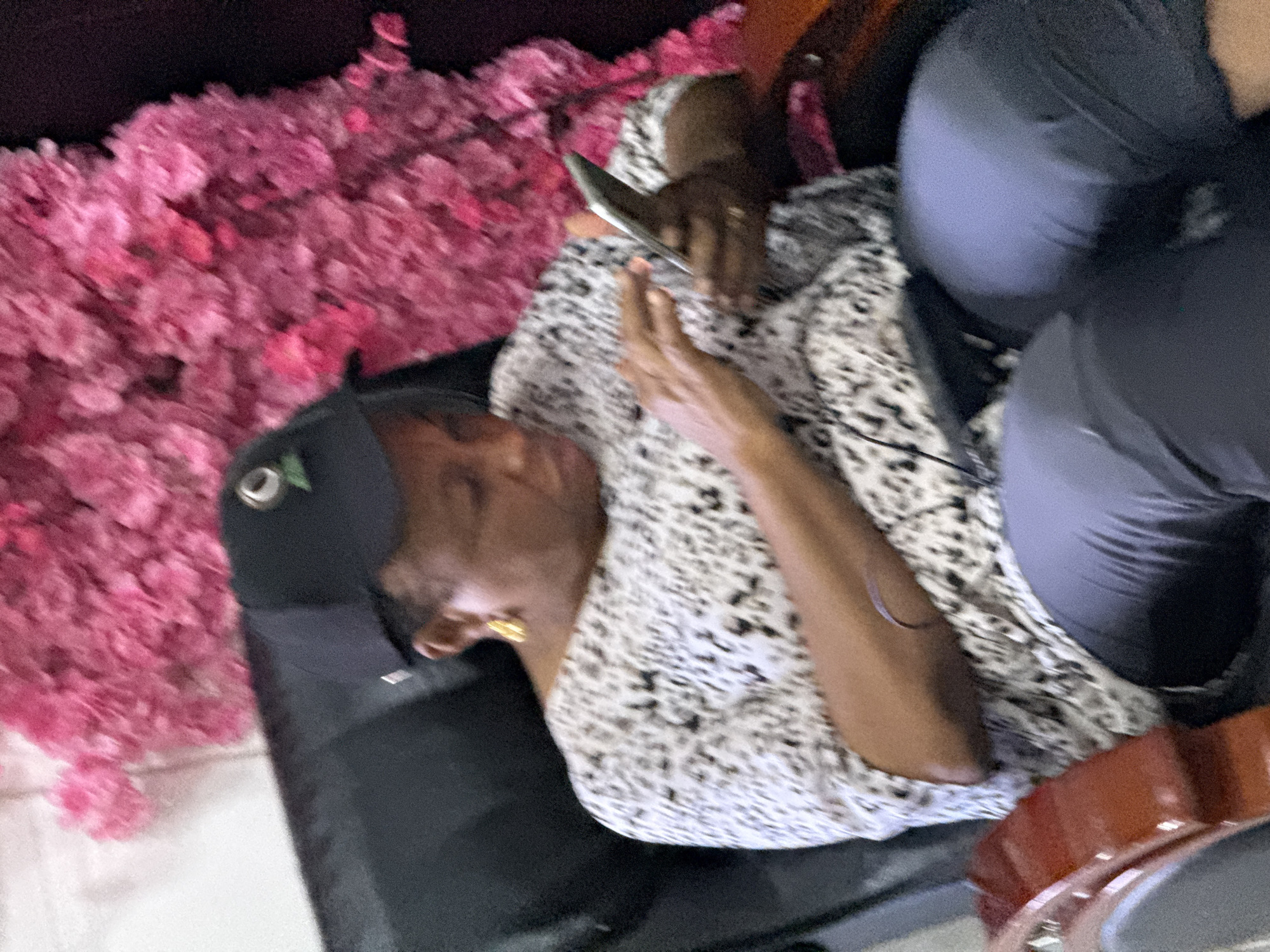 A middle-aged woman wearing a black baseball cap and a patterned white and black shirt is seated on a dark-colored surface, possibly a chair or sofa. She is holding and looking at a mobile phone. A mass of pink flowers is positioned to her left. Another person, likely wearing gray or black pants, is partially visible to the woman's right. The setting appears to be indoors, but further detail to identify the location within Lugbe, Nigeria, is not available.