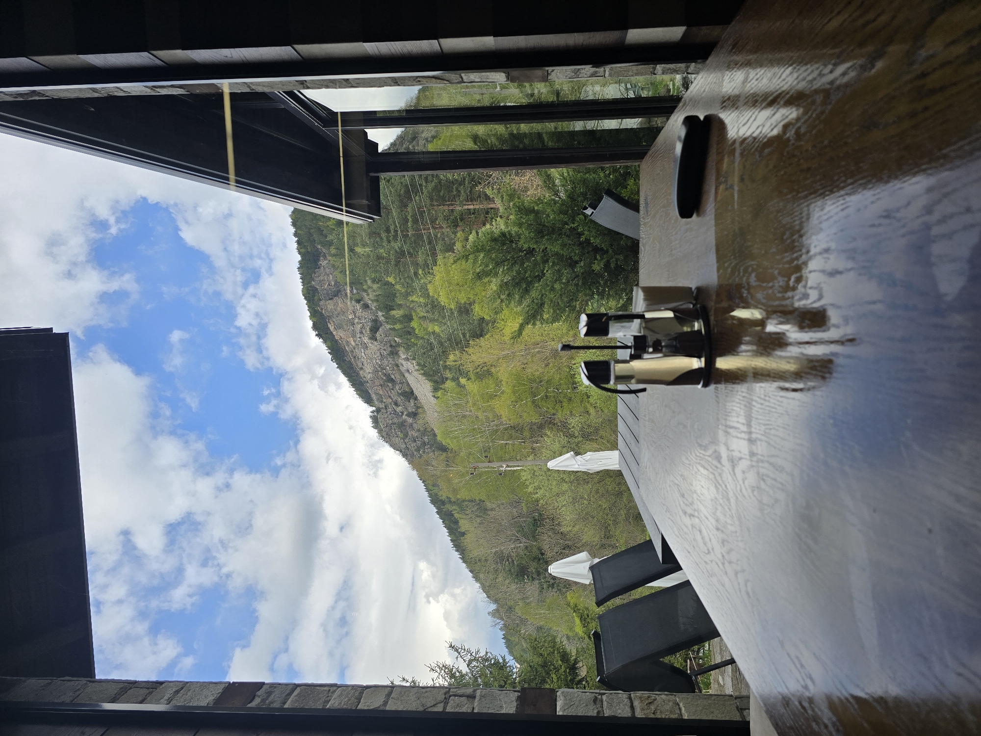 A slightly rotated indoor shot shows a wooden table with some objects on it, with a view of a mountain landscape through a window or opening.
On the table are two small metal objects, possibly part of a device, and a black object, maybe a smartphone or remote. The table appears to be polished and reflects some of the outdoor scenery.
Outside, a mountain range covered with green trees and some bare patches is visible. The sky is blue with some white clouds. There are also some power lines visible in the distance. The location is Sapareva Banya, Bulgaria. The presence of an outdoor table and mountain view implies a possible seating area on the building's porch or a terrace.