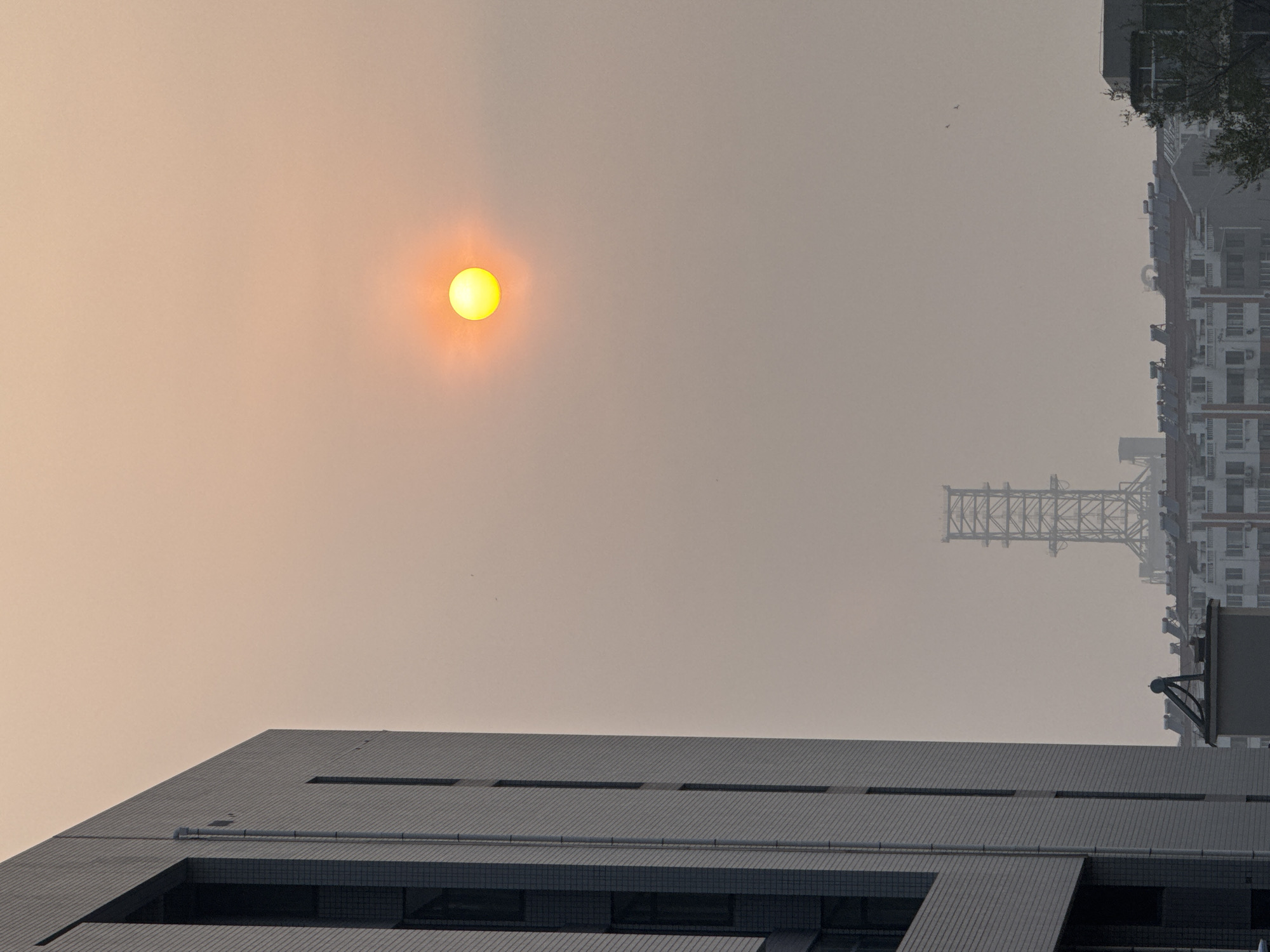 The image depicts a hazy, overcast scene, possibly in Zhongtun, China, judging by the architectural style. In the sky, a blurred, pale yellow sun is visible. Below, there is a modern building with a flat, segmented roof. Beyond that, a lattice tower stands, enveloped in the haze, next to a densely populated residential building. The perspective is tilted.