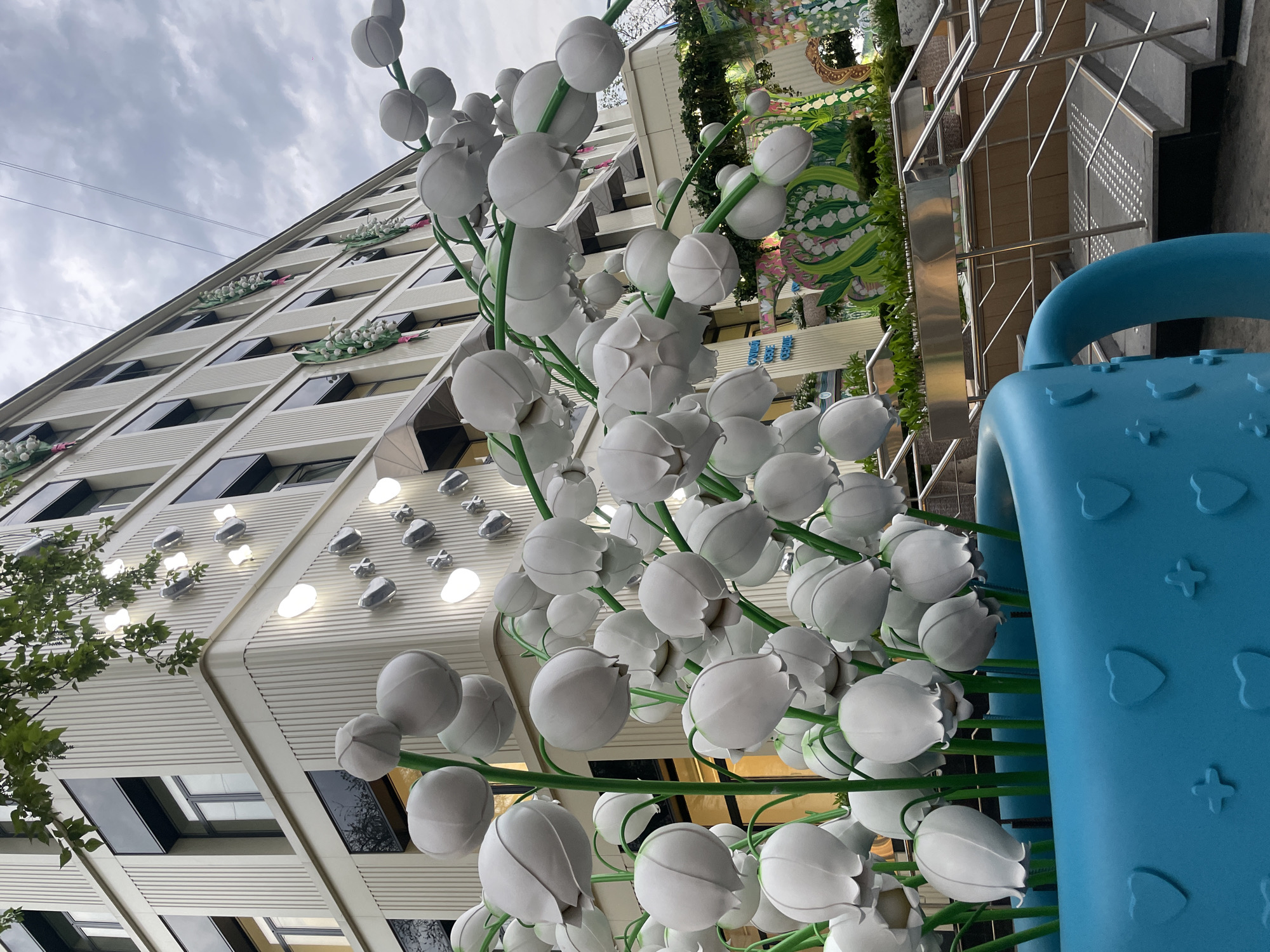 A low-angle outdoor shot depicts white, ball-shaped floral sculptures with green stems in the foreground, set against a backdrop of a modern building with geometric architectural elements and a sky filled with overcast clouds. The scene includes elements suggesting the location is in Moscow, Russia, with some details of building decorations. To the right, a blue, plastic object with embossed heart and flower shapes is partially visible. The perspective is skewed, suggesting the photo was taken at an unusual angle, looking up at the structures.