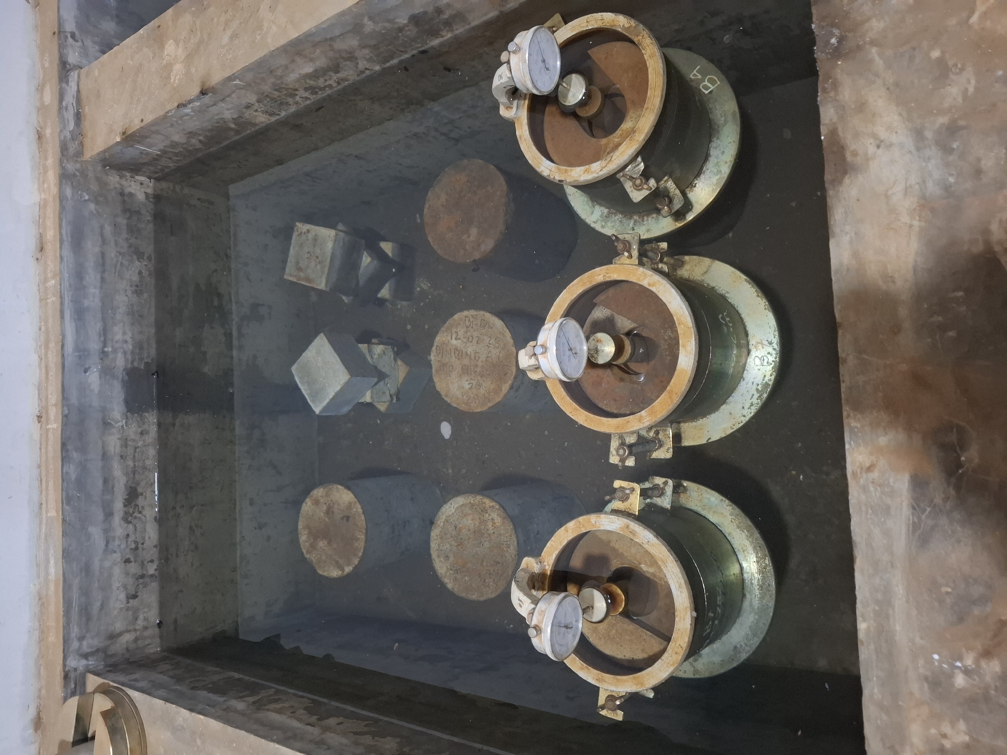 A concrete testing laboratory in Kutasirna, Indonesia, houses a water tank filled with murky water containing various concrete test specimens. Submerged in the water are several cylindrical concrete samples and some cube-shaped concrete samples. In the foreground, three cylindrical metal containers, each topped with a dial gauge, are partially submerged. These are likely part of a test setup, possibly for measuring expansion or contraction of the concrete samples. The water tank itself appears to be constructed of concrete and shows signs of age and use. The overall environment suggests a functional but not pristine laboratory setting.