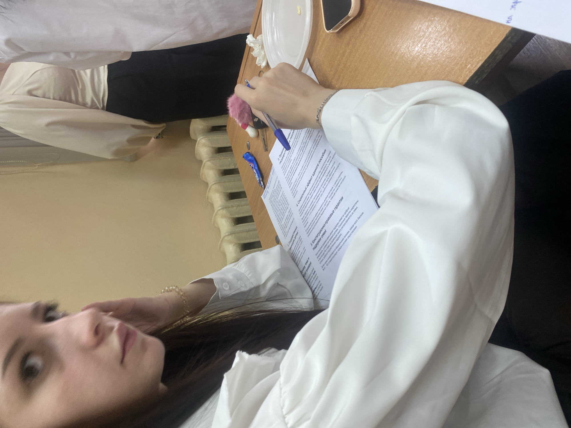 A young woman with long dark hair, wearing a white blouse with puffed sleeves and a gold bracelet, is seated at a wooden desk. She is looking to her left, with her left hand touching her face. On the desk, in front of her, is an open document with text in Russian. A blue pen is in her right hand, poised over the document. Other objects on the desk include a small pink fuzzy item, a key, a coin, a blue wrapper, a white paper plate, and a smartphone. The background shows a radiator and a wall. The scene appears to be indoors, possibly in a classroom or office. There is no specific indication of the location being in Dyakovo, Russia.