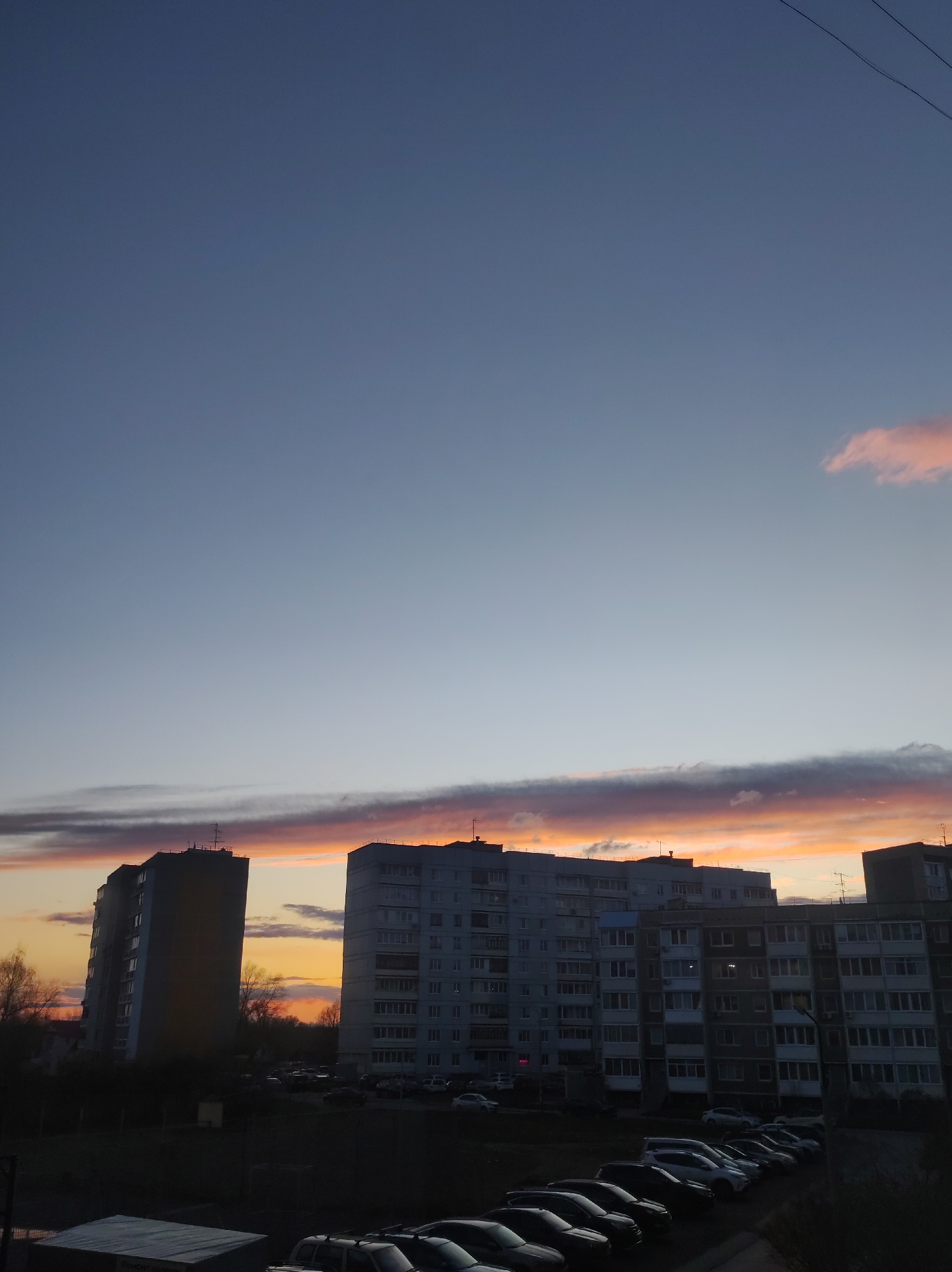 The image captures an urban residential scene in Chaikovsky, Russia, during sunset or dusk. Multi-story apartment buildings, characteristic of Soviet-era architecture, dominate the lower half of the frame. A taller, narrower building stands on the left, while a wider, longer structure occupies the center and right, both silhouetted against the light. Antennas are visible on their rooftops.
The sky is a prominent feature, transitioning from a clear, deep blue-gray at the top to a vibrant display of colors near the horizon. A long, horizontal band of clouds is brightly illuminated by the setting sun, showcasing warm orange, pink, and purple hues. A smaller, isolated pink cloud is visible in the upper right, and faint overhead wires stretch across the sky.
In the mid-ground and foreground, numerous cars are parked in rows, indicating a residential parking lot. Leafless trees are visible between the buildings and against the horizon, suggesting late autumn or winter. The ground and buildings are cast in deep shadow, emphasizing the dramatic colors of the sunset. No human activity or discernible text is visible.