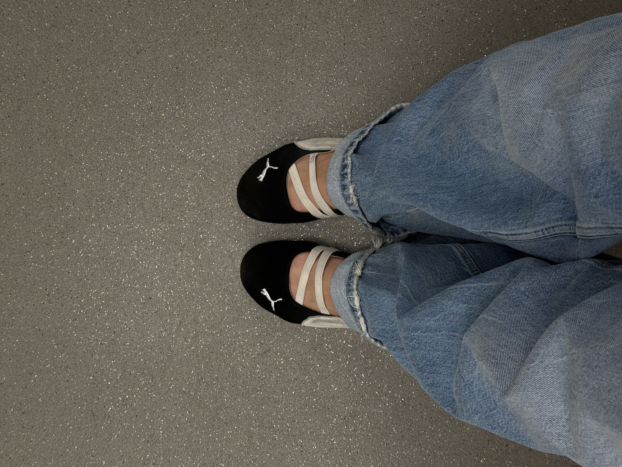 The image, captured from a downward perspective, displays the lower legs and feet of a person. The individual is wearing light blue, wide-legged denim jeans that are casually draped or slightly cuffed at the ankles, showing some wear and creasing. On their feet are black ballet flat-style shoes, which feature three cream-colored elastic straps extending across the instep. A distinctive white Puma logo, depicting a leaping puma, is prominently visible on the toe area of both shoes.
The person's feet are positioned on a gray, speckled floor surface. This floor appears to be a composite material, possibly terrazzo or quartz, characterized by numerous small, reflective flecks and darker grains embedded within a lighter gray base, creating a textured visual effect. The scene suggests an indoor public or commercial setting. The activity depicted is static, with the person's feet resting on the floor. Based on the provided context, the location is Moscow, Russia.