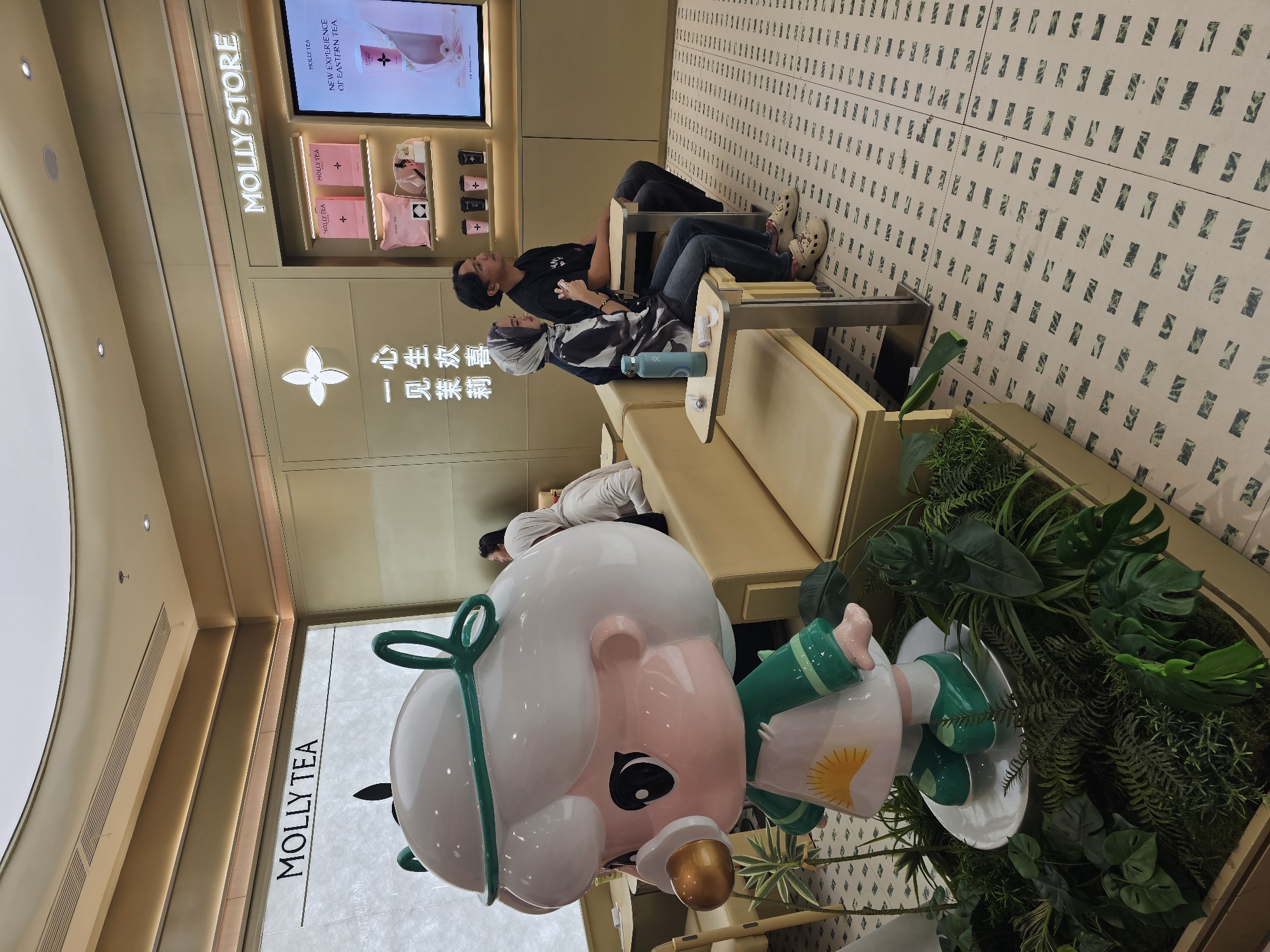 This is a medium shot photograph of the interior of a "Molly Tea" store, likely located in the city of Condongcatur, Indonesia, given the context. In the foreground, to the left, is a large, colorful statue of a cartoon character with white hair, green trim, and a sunburst on its apron. The statue stands amidst artificial plants. To the right of the statue, a beige couch is visible, with two people seated on it. A man in a black t-shirt and dark pants is on the right, and a woman in a patterned headscarf and black and white striped shirt is to his left. Both are looking forward, seemingly at a display on the wall. On the wall behind them, there is signage that reads "MOLLY STORE" and "MOLLY TEA". A digital screen displays advertisements for "Molly Tea" products. The floor is tiled with a light-colored background and a repeating pattern of dark rectangles. The lighting is soft and ambient, originating from recessed ceiling lights and accent lighting above the displays.