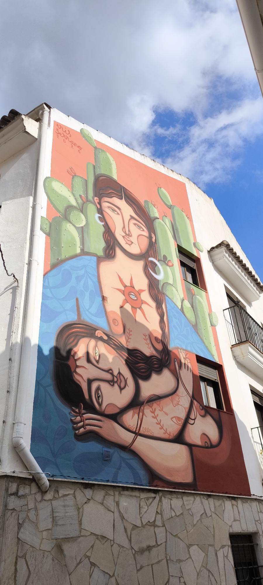 A vibrant mural adorns the side of a white building in Fanzara, Spain, depicting two stylized female figures against a backdrop of large green cacti and a bright blue sky with white clouds. The upper figure has long braided dark hair, large hoop earrings, and a sun-like symbol with an eye at its center on her chest. She is positioned behind and above the lower figure. The lower figure, with dark hair and green eyes, is lying down, her head resting on her arm, with intricate vine-like tattoos on her skin. Both figures have a flattened, graphic artistic style. The building itself has a white facade, with a tiled roof visible at the top. A white downspout runs along the left side of the building, and a small ventilation grate is visible on the wall within the mural's blue background. The lower part of the building is faced with irregular light-colored stones. A window with dark shutters and a black metal railing on a balcony are visible on the right side of the building, partially superimposed by the mural. The mural is signed "Pus Serrano 2026" in red lettering in the upper left corner.