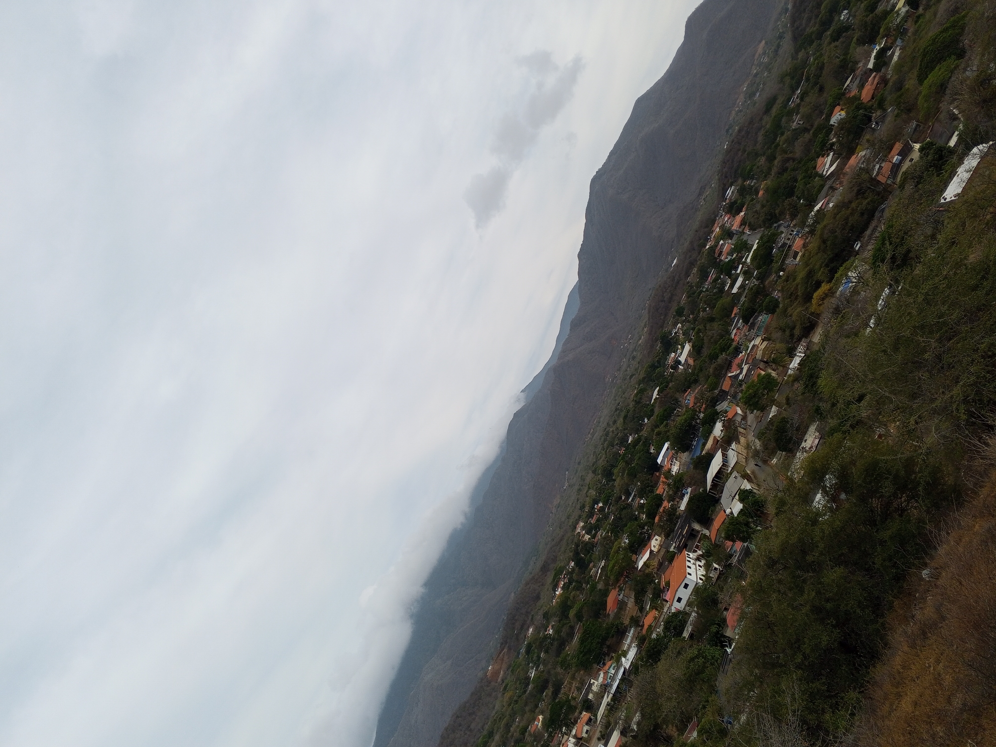 The image shows a view of a settlement on a mountainside, likely taken on a cloudy day. The settlement is comprised of densely packed buildings, many with red tile roofs, interspersed with trees and vegetation. To the left, the mountainside rises sharply, partially obscured by clouds or fog. The overall perspective is tilted, giving a sense of height or an elevated viewpoint. The location is Unknown, Unknown.