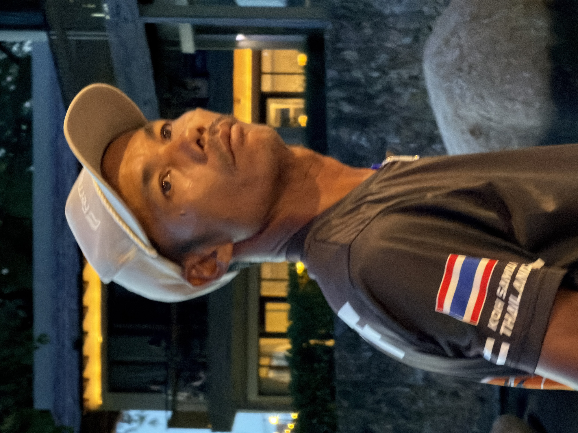 The image captures a close-up profile of a man wearing a light-colored baseball cap and a dark t-shirt with the Thai flag and "KOH SAMUI THAILAND" printed on the sleeve. He appears to be standing outdoors in Baan Bo Phut, Thailand, possibly near a building with lit windows in the background and some foliage. The lighting suggests it might be evening. The man's gaze is directed to his right.