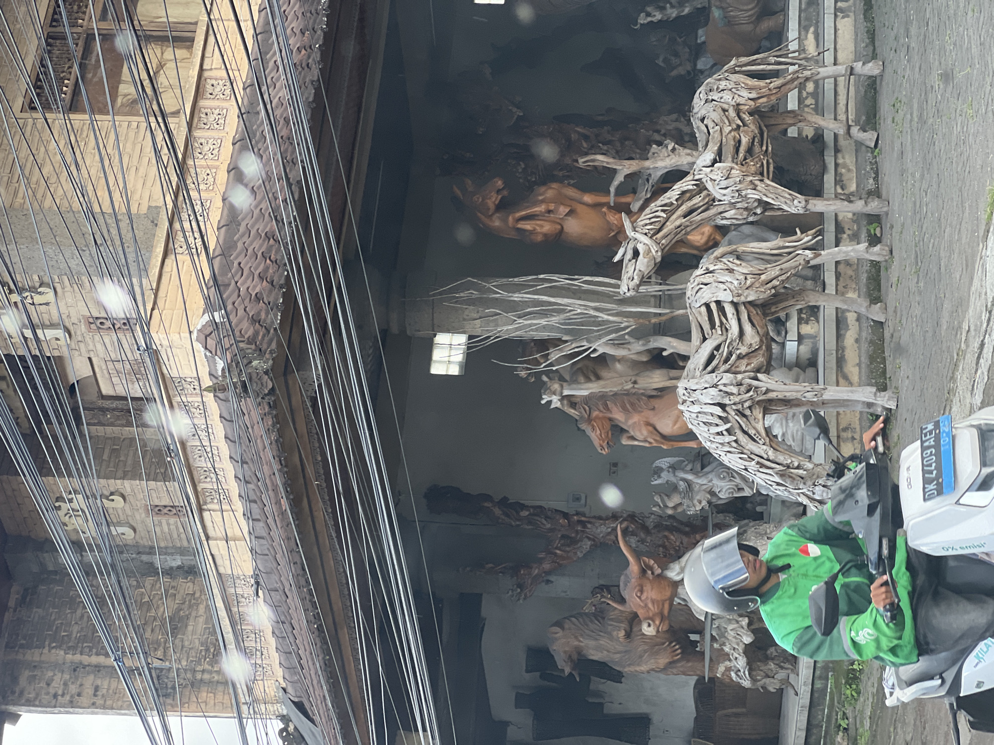 The image shows a street scene in Ubud District, Indonesia, focused on a shop selling wooden sculptures. The storefront displays a variety of wooden animal sculptures, including horses, deer, and a bull, in both natural wood tones and a lighter, driftwood-like finish. A person wearing a green jacket and helmet is sitting on a scooter in front of the shop, facing the camera, partially obstructing the view of the sculptures. The scooter has a license plate visible, reading "DK 4409 AEH". Overhead, numerous electrical wires run across the scene, obscuring a portion of the building's facade, which features decorative carvings and a tiled roof. The overall impression is a blend of local craftsmanship and everyday street life in Ubud.