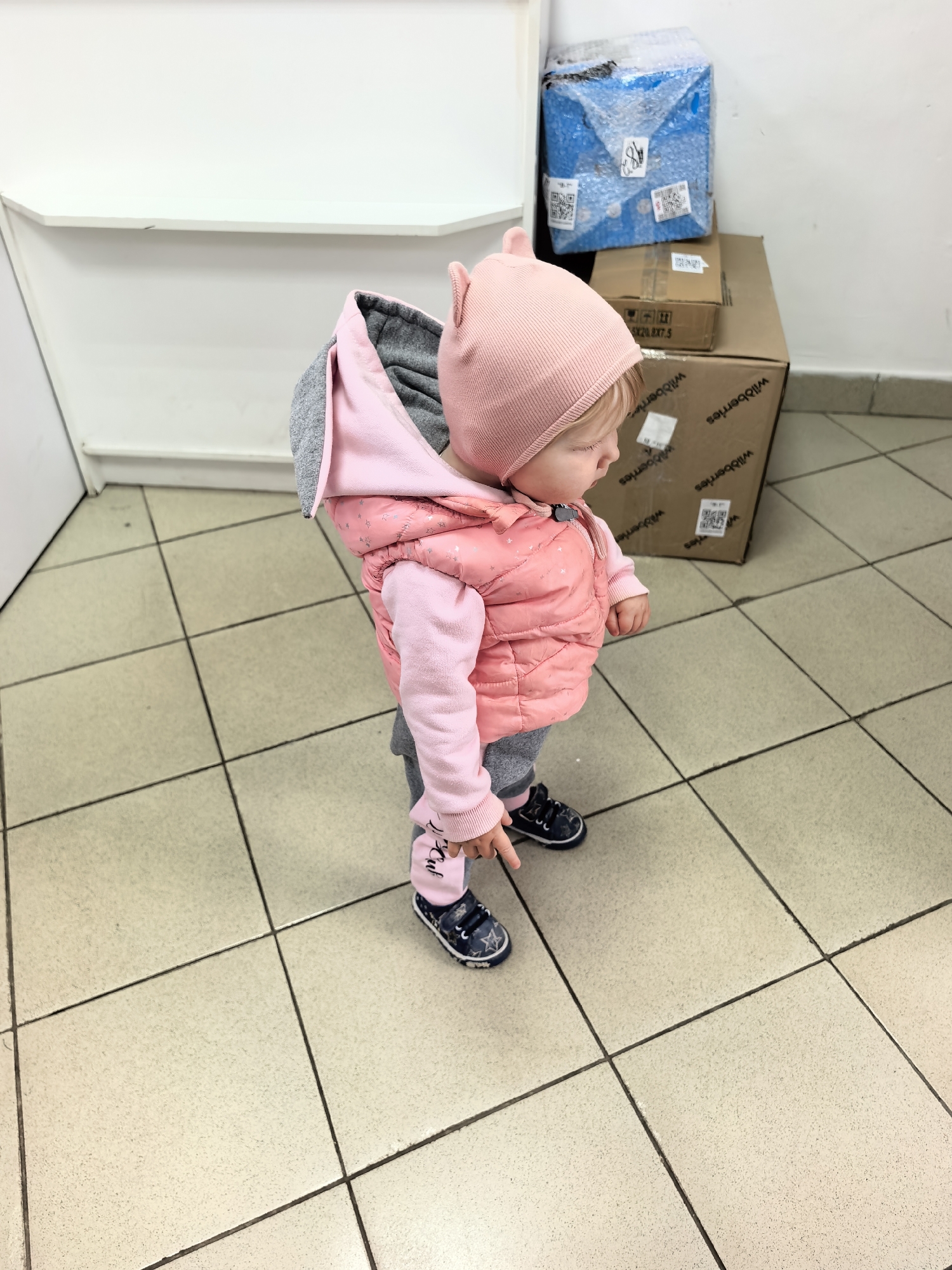 In Sterlitamak, Russia, a young toddler, likely a girl based on her attire, stands on a tiled floor in what appears to be a delivery or waiting area, possibly a post office or parcel pickup point. The toddler is dressed in a pink hooded jacket adorned with subtle star patterns, a pink beanie with cat ears, and grey sweatpants. She is wearing dark blue velcro shoes with white star accents. Her right hand is extended, pointing downwards towards the floor. In the background, several cardboard boxes are stacked, some wrapped in plastic or bubble wrap. One prominently visible box features the "wildberries" logo. The environment is sterile, with white walls and a white shelf above. The lighting is even, suggesting an indoor setting.