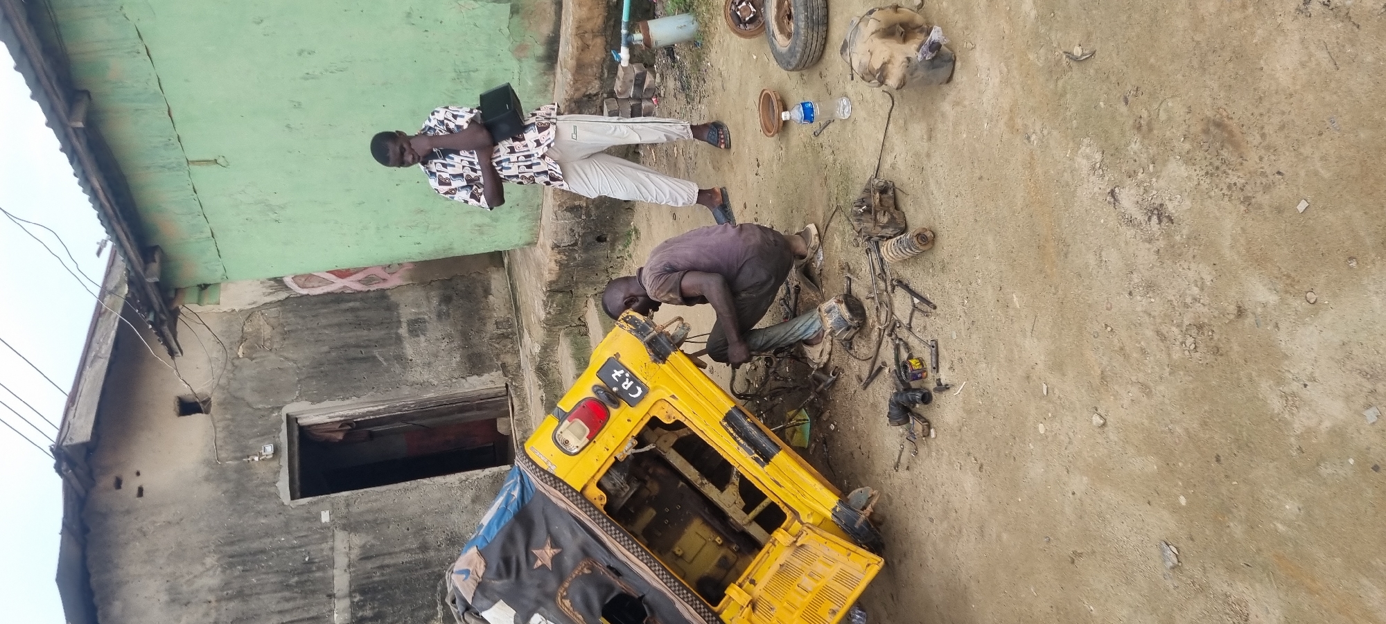 In the city of Ikorodu, Nigeria, on a dirt ground, a yellow auto rickshaw is being repaired. The auto rickshaw is tilted on its side and parts of its engine are exposed. A man, dressed in a dark brown t-shirt and grey pants, is crouched down and working on the engine. Another man, dressed in a white shirt with a multicolored pattern and white pants, stands a few feet away, observing. Scattered around the auto rickshaw are various tools, car parts, and a water bottle. Behind the scene, there is a dilapidated building with a green and grey exterior. The sky is overcast.