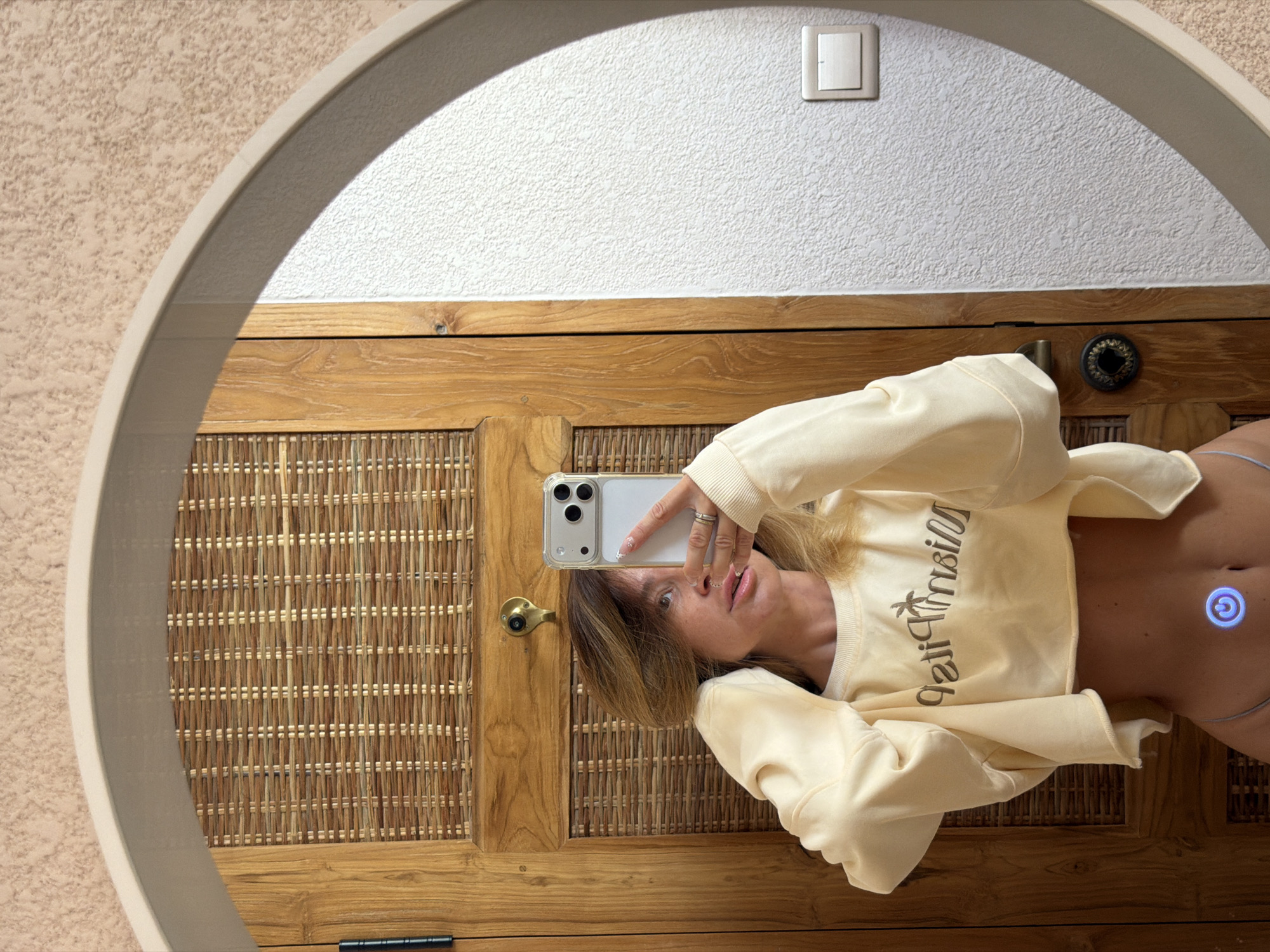 A mirrored selfie of a woman is reflected in a round mirror, taken indoors. The woman is holding a smartphone, a white iPhone, in her right hand to capture the image. She is wearing a pale yellow crop top with lettering. The background features a cabinet with a woven front, wooden frame, and handles. Above the cabinet, the wall is white with a light switch. The reflection in the mirror also shows an adjacent wall with a textured beige surface. The location may be in Mengwi, Indonesia.