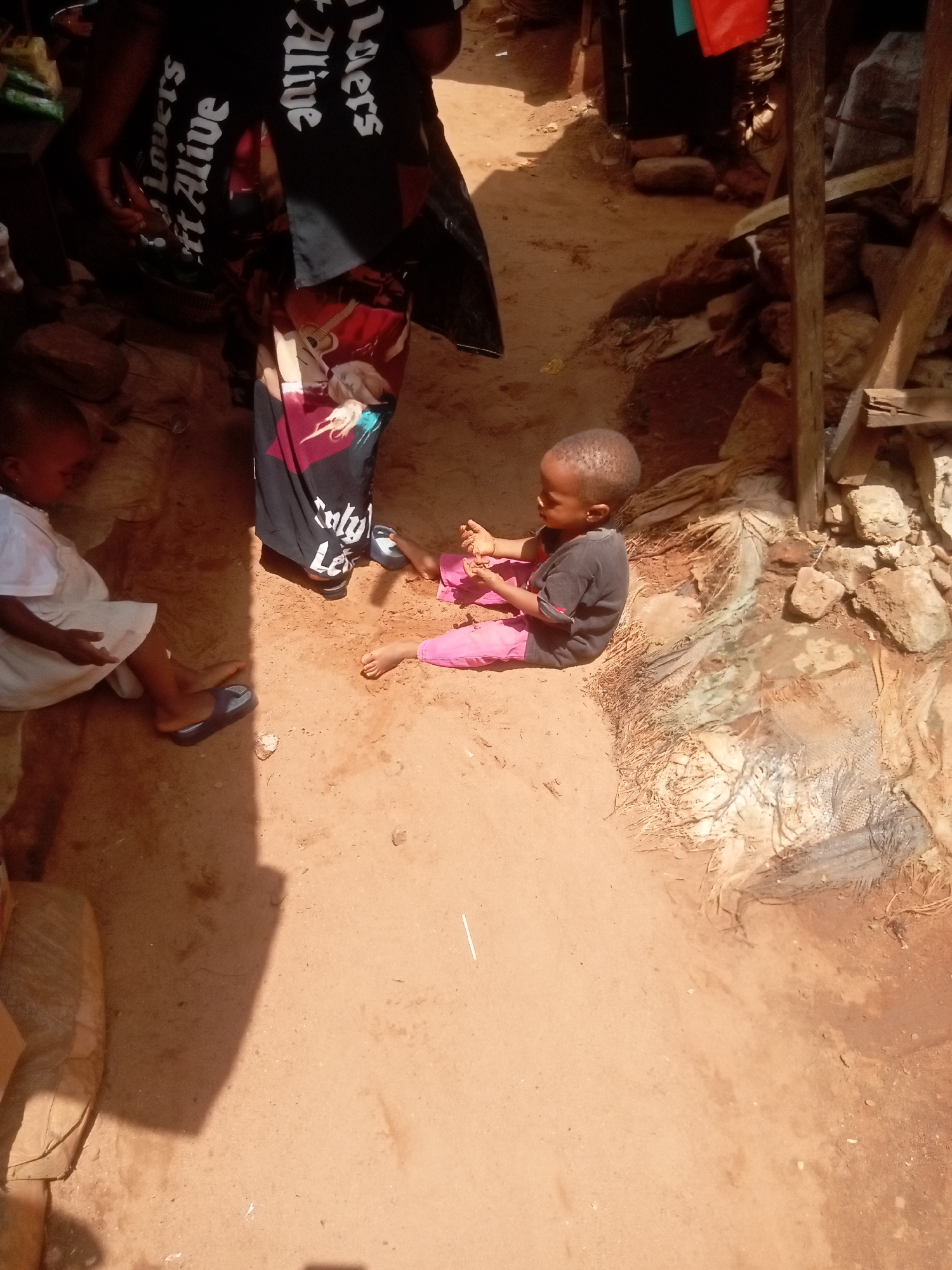 The scene depicts two children and an adult in Ogidi, Nigeria. One child, wearing a white dress and blue shoes, sits on the bare earth to the left of the frame. Another child, in a black shirt and pink pants, sits on the earth in the center of the frame, appearing to play with small objects in their hands. An adult stands behind them, wearing a dark top with the words "Lovers Alive" printed on it, and a patterned skirt. The ground is predominantly sandy soil with patches of rock and sparse vegetation. Part of a wooden structure is visible on the right. The light suggests it is daytime, with strong shadows indicating a sunny environment.