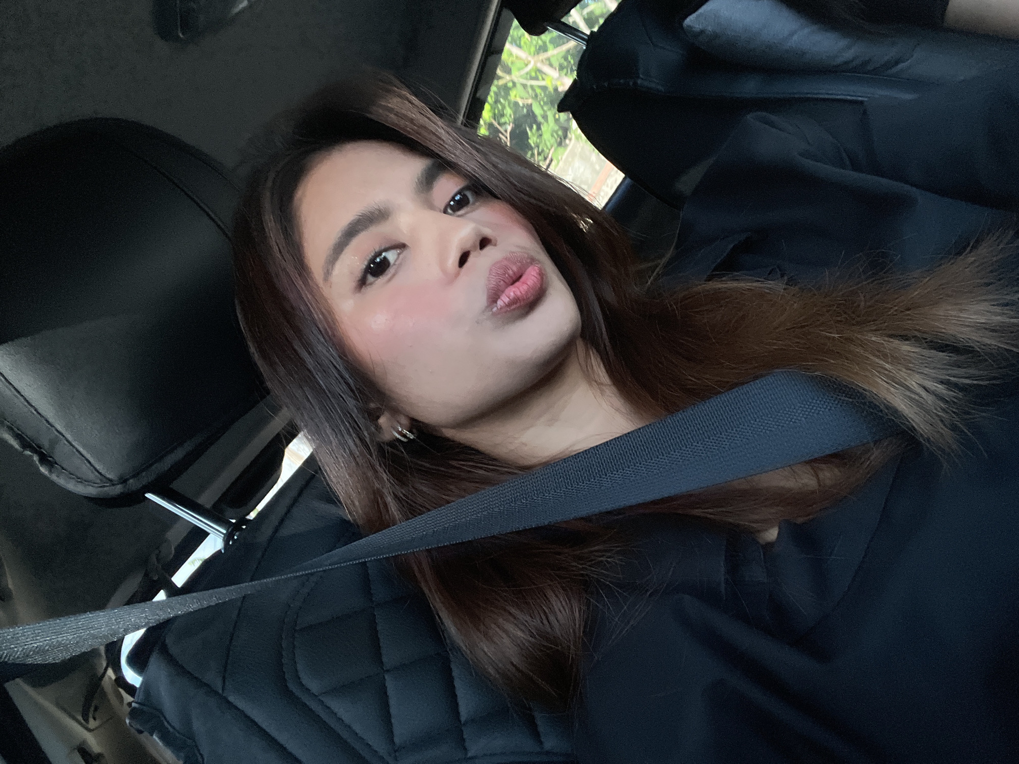 A female individual is seated in the front passenger seat of a vehicle, looking directly at the camera with a pouting expression. She has long, dark brown hair, styled with a side part, and is wearing a dark, collared shirt. A seatbelt is fastened diagonally across her left shoulder and chest. Her face exhibits applied makeup, including blush on her cheeks, eyeshadow, and lip color. A small silver hoop earring is visible on her left ear. The vehicle's interior features dark-colored upholstery, with a quilted pattern visible on the back of the driver's seat. A headrest is present in the upper background, featuring a metallic accessory attached to its support post. Through the window on the right side of the image, green foliage from trees and bushes is visible, suggesting an outdoor, vegetated environment. A light-colored structure, possibly concrete, is partially observable behind the greenery. The scene is located in Davao City, Philippines.