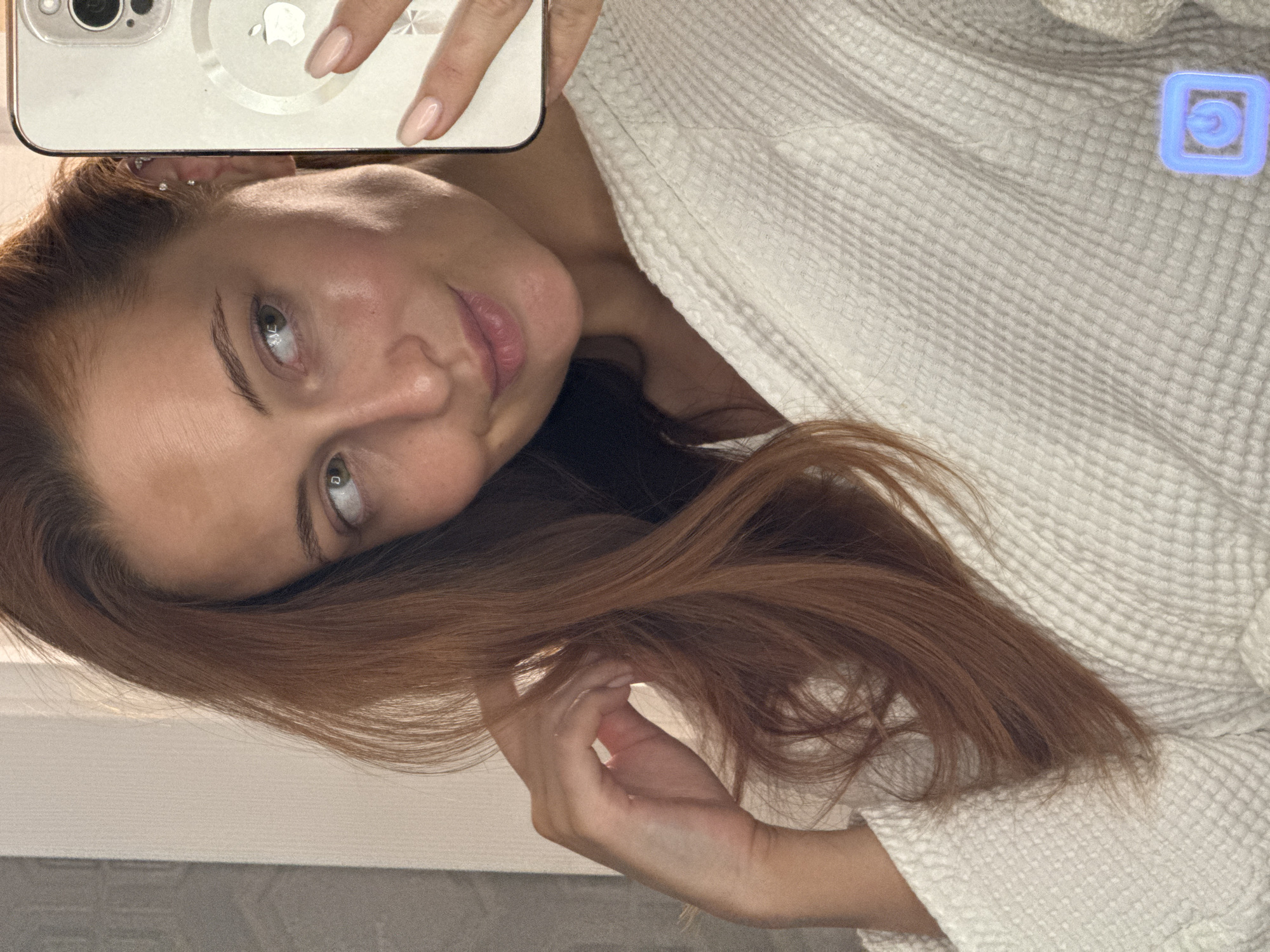 A selfie taken by a light-skinned woman with auburn hair. She is wearing a white robe and holding a smartphone with a clear case. The location is indeterminate, and there's no evidence to suggest that this image was taken in Chaikovsky, Russia.