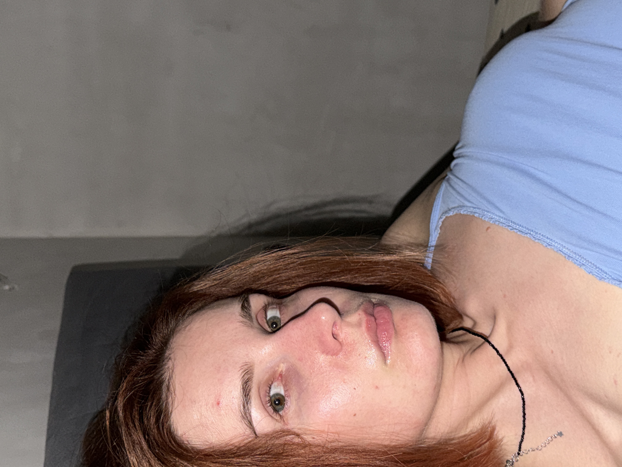 The image is a selfie taken from a low angle, showing a woman lying down. She has reddish-brown hair, fair skin, and green eyes. She is wearing a light blue tank top. A thin black necklace with a small silver charm is visible around her neck. The background includes a grey wall and a grey surface, possibly a headboard or bed frame. The setting is undetermined and the location in the city of Chaikovsky, Russia is unsupported by the image.