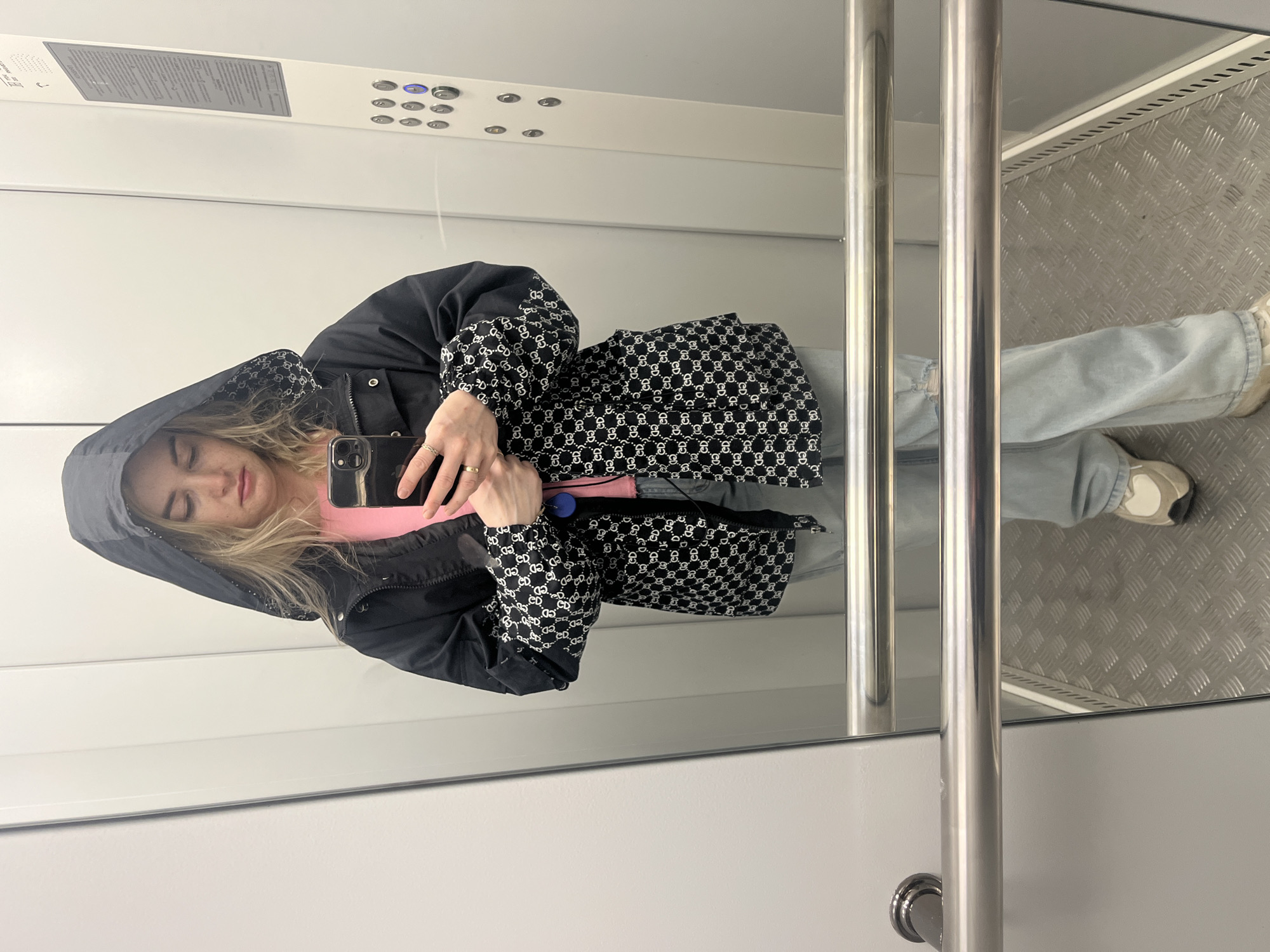 A female is taking a selfie in an elevator. She is wearing a black hooded jacket with a repeating "GG" logo pattern, a pink shirt, and light blue jeans. The elevator has white walls, a control panel with buttons at the top, and a metal floor. The location is Chaikovsky, Russia.
