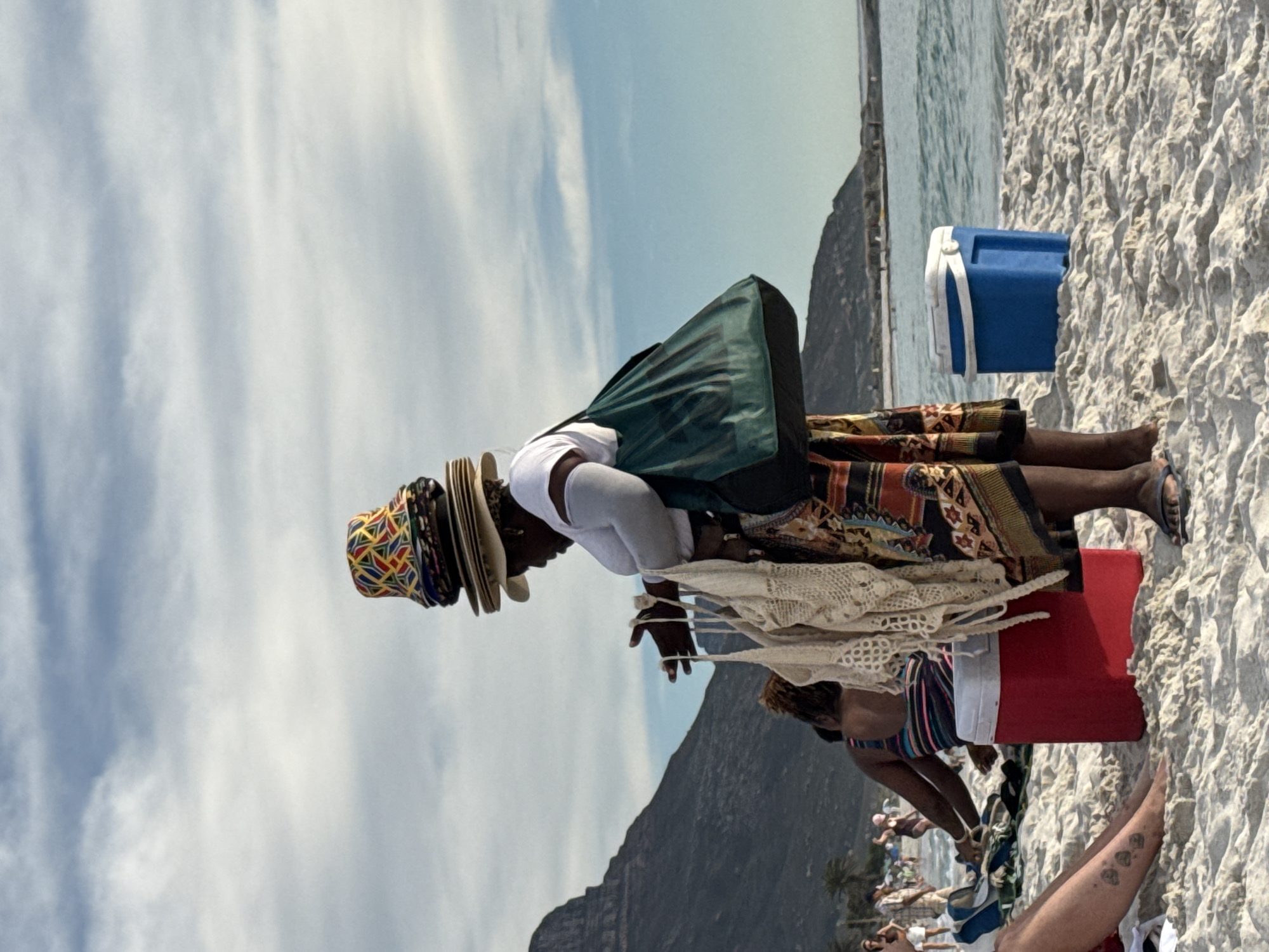 The image shows a beach scene in Cape Town, South Africa. A woman is standing on the sand, selling hats. She is wearing a white shirt, dark skirt, and multiple hats stacked on her head. She has a green bag on her back, and she is holding up a white crochet item, possibly for sale. Next to her, a cooler is on the sand. Behind her, people are enjoying the beach, and a mountain is visible in the background. The sky is blue with some clouds.