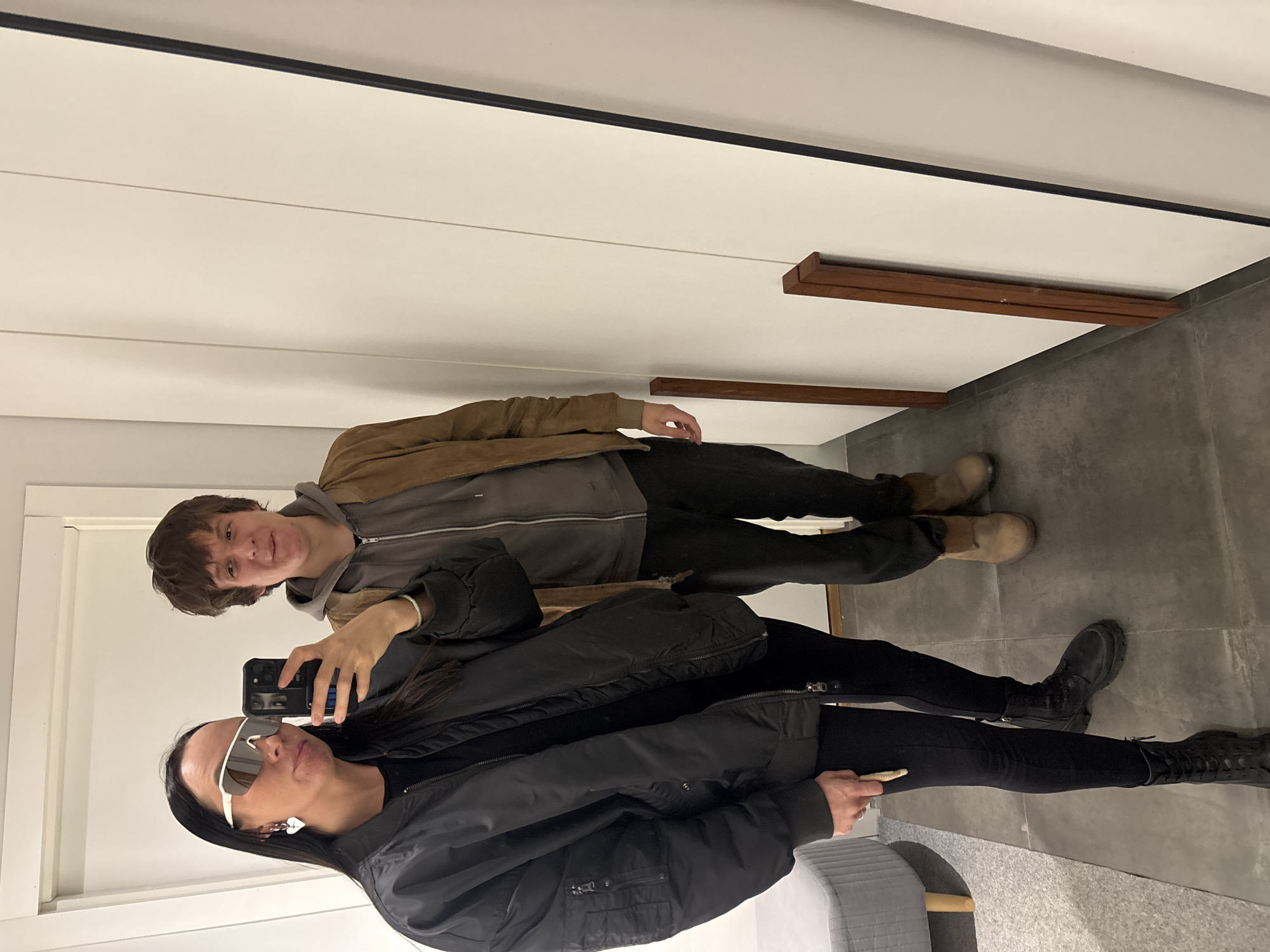 A female is depicted taking a mirror selfie with a male companion in an indoor setting. The female, on the left, has long dark hair, wears white-framed sunglasses, a silver heart earring on her left ear, a black bomber jacket, a black undershirt, black leggings, and black combat boots. She holds a black smartphone in her right hand to capture the image and a small, light-colored elongated object in her left hand, which rests on her left thigh. The male, positioned to her right, has brown hair and wears a brown suede jacket over a grey hooded sweatshirt, dark trousers, and light brown boots. Both individuals are looking towards the camera. The environment features plain white paneled walls and a grey tiled floor. A large mirror with a dark frame occupies the right side of the scene, reflecting the subjects. A grey upholstered cylindrical pouf with wooden legs is partially visible on the far left. Two slender brown wooden strips lean against the wall near the floor on the right. The specific location within Chaikovsky, Russia, is not ascertainable from the visual evidence.