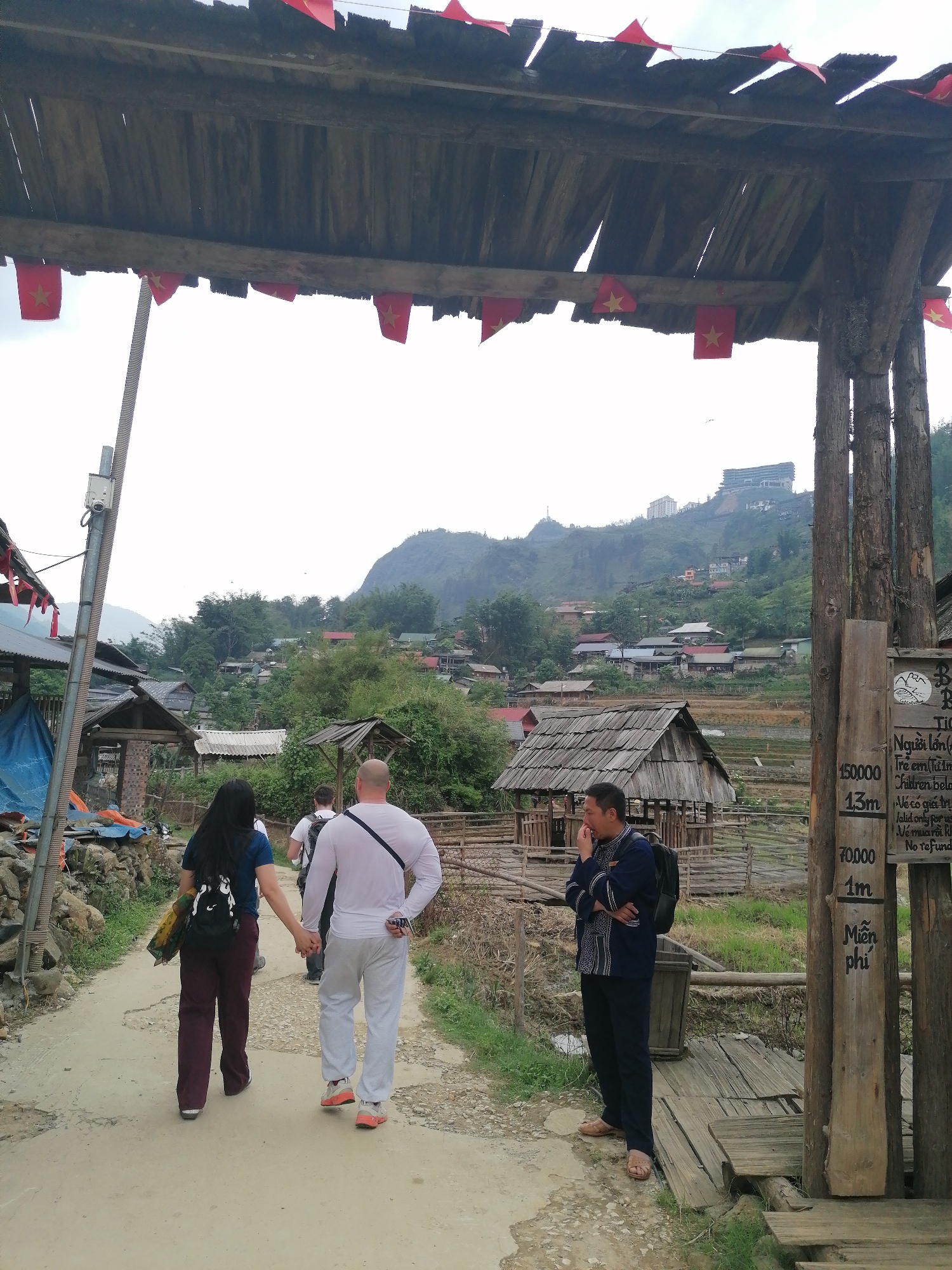The image depicts a vibrant scene at what appears to be the entrance to Cat Cat Village in Vietnam. A rustic wooden gate, adorned with numerous small red flags bearing a yellow star, frames the top of the photograph. Beneath it, a dirt path leads into the village, winding past traditional wooden structures and terraced fields.
In the foreground, four individuals are visible. A couple walks away from the camera, holding hands, suggesting they are visitors or tourists exploring the area. The woman wears a dark top and maroon pants with a backpack, while the bald man beside her wears a white long-sleeved shirt and light grey sweatpants, also carrying a backpack. Behind them, another person is partially obscured. To the right, near a wooden signpost, a man in a distinct dark blue traditional-patterned shirt and dark pants stands, looking pensive or thoughtful with his hand near his mouth. He also carries a backpack.
The setting combines traditional village elements with distant signs of modernity. Beyond the immediate village structures and lush green hillsides, a large, modern building, possibly a hotel or resort, can be seen on a distant hilltop, indicating the area's development. The sky is bright but overcast, suggesting daytime with diffused light. A power pole with a CCTV camera is visible on the left.
A prominent wooden signpost on the right details entrance fees. The visible text, partially in Vietnamese and English, states: "Người lớn" (Adults) 150,000 (VND), "Trẻ em" (Children below) 1.3m pay 70,000 (VND), and those below 1m enter "Miễn phí" (Free). It also mentions "Vé có giá trị" (Ticket valid) and "No refund," indicating a ticketing system for entry into the village or a specific attraction within it.