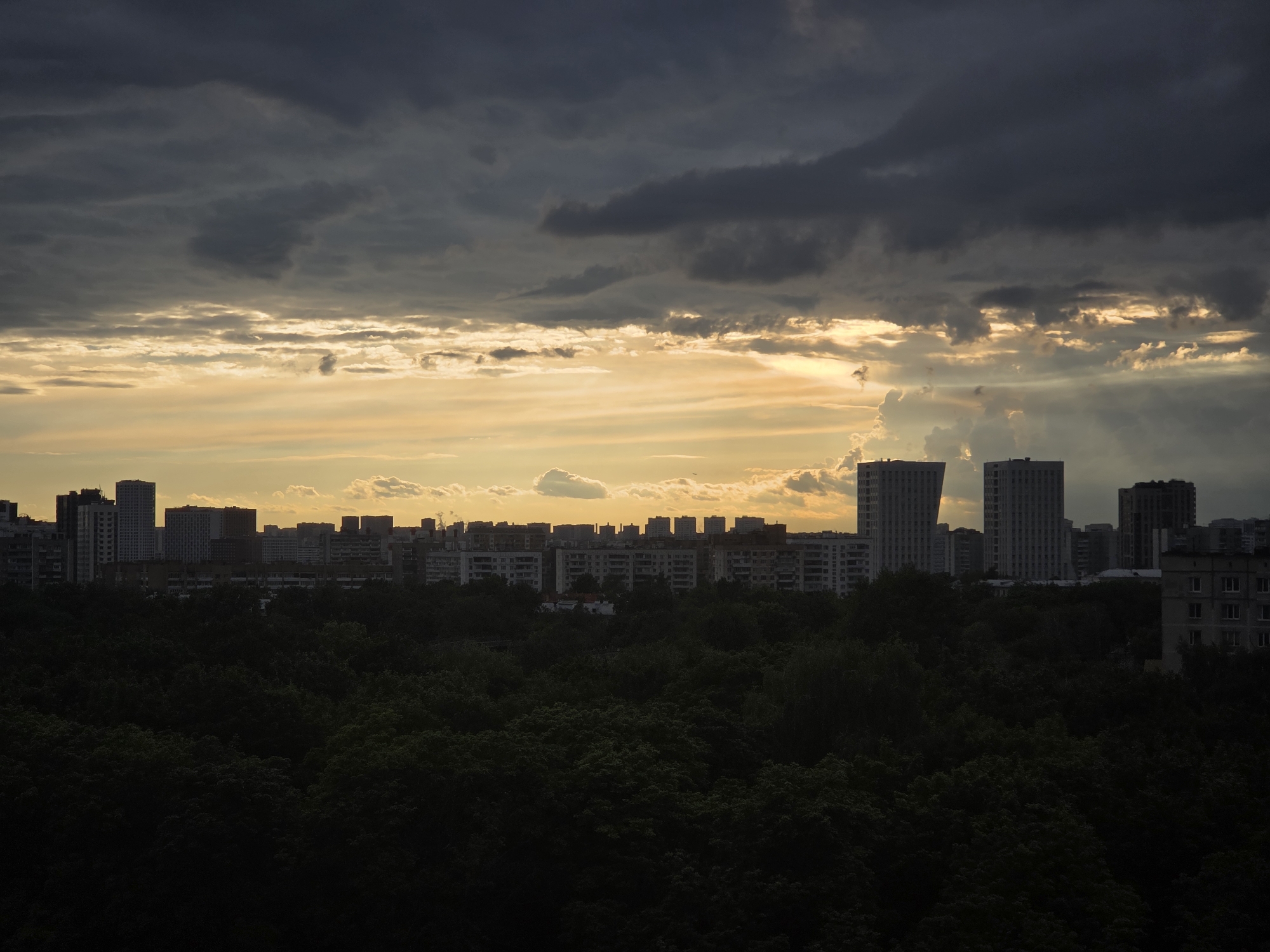 This media file shows a city skyline at dusk, with dark clouds overhead contrasting with a golden sunset. The scene depicts a dense canopy of trees in the foreground, obscuring much of the lower buildings. Behind the trees, a line of apartment buildings and taller structures stretches across the horizon. The sky is dramatic, with patches of bright light breaking through the dark, ominous clouds, suggesting an approaching storm or the remnants of one. The overall mood is somber and atmospheric. The time of day is clearly evening, as indicated by the sunset colors and the darkening landscape. The location is Moscow, Russia, as confirmed by the architectural style of the buildings, which are characteristic of Soviet-era housing blocks and more modern high-rises. No people or specific objects are discernible in the foreground or midground. There is no visible text in the image.