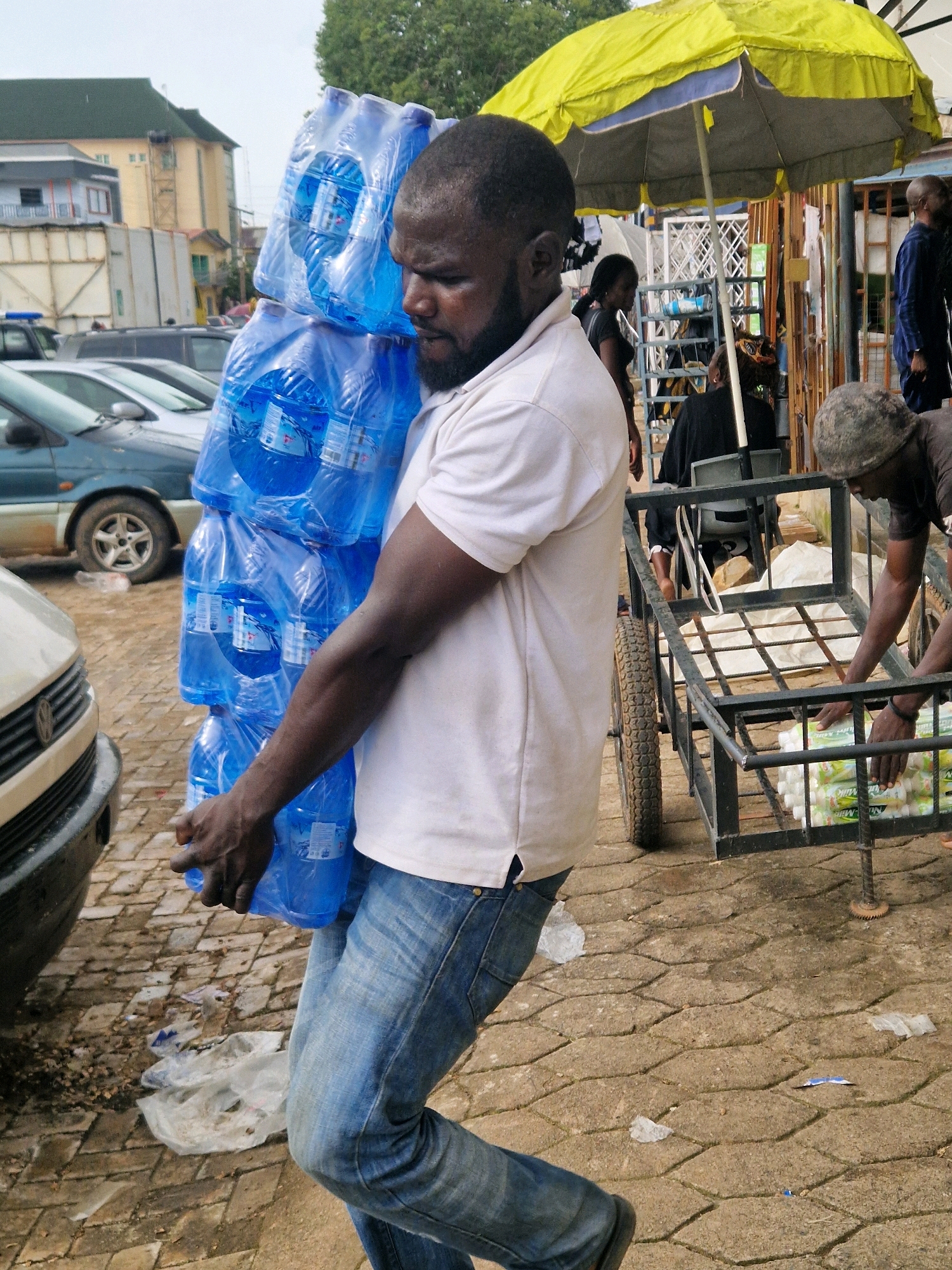 A man is carrying a large stack of bottled water in the city of Jos, Nigeria. He is wearing a white t-shirt and blue jeans. The bottled water is stacked high on his shoulder and chest, with the bottles enclosed in plastic wrapping. In the background, there is a yellow and blue umbrella, a cart with some items, and other people. The ground is paved with interlocking stones. The overall scene suggests a commercial area, possibly a street market, where goods are being transported.