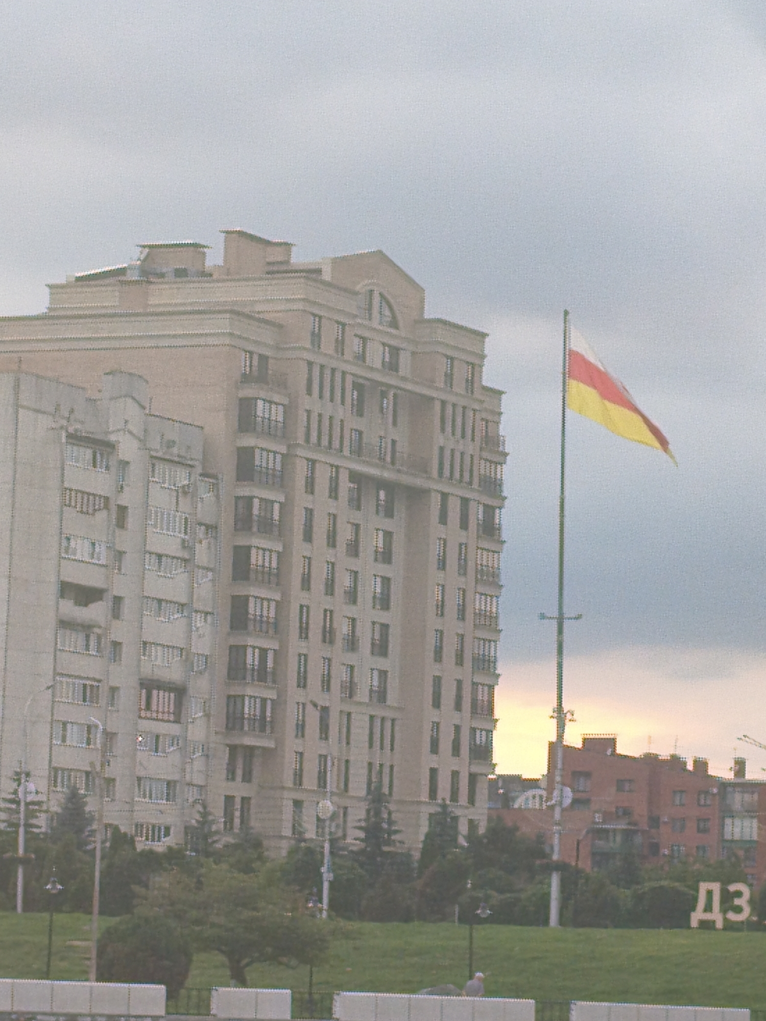 A wide shot captures a cloudy sky over a cityscape featuring several large apartment buildings. A tall flagpole with a tricolor flag, yellow, white, and red, stands prominently in the foreground. The flag appears to be fluttering in the breeze. One of the apartment buildings is a light-colored, modern structure with numerous windows and balconies. To its left, another, older apartment building is visible, also with many windows. In the background, a reddish-brown building with a patterned facade is seen, and a portion of a yellow sky can be observed peeking through the clouds, suggesting it may be sunrise or sunset. Large, white letters spelling out "ДЗ" are visible on a green grassy area in the lower right portion of the frame. Trees and lampposts dot the landscape between the buildings and the foreground. The image appears to be taken from a slightly elevated perspective, looking across a green expanse.