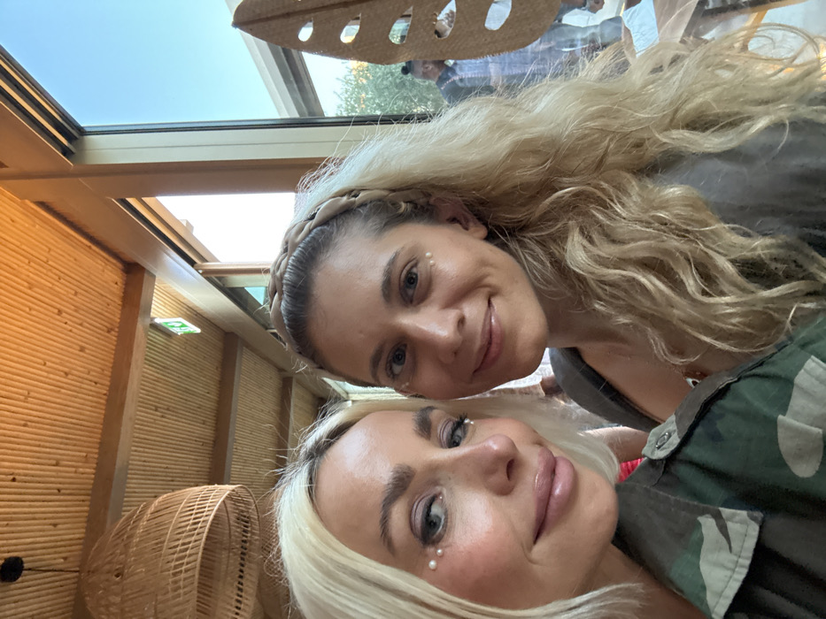 The image displays two adult females in a close-up, selfie-style photograph taken indoors at a venue in Dubai, United Arab Emirates. The female on the left has blonde hair, light-colored eyes, prominent lips with gloss, and wears a camouflage-patterned jacket. She has three small pearl-like embellishments affixed below her left eye and one below her right. The female on the right has long, wavy blonde hair, wears a tan braided headband, and has one small pearl-like embellishment affixed below each eye. Both individuals are looking towards the camera with faint smiles.
The indoor environment features a ceiling constructed from light-colored wooden slats supported by wooden beams, from which a large, woven wicker pendant lamp hangs in the upper left. A green emergency exit sign with a white running man pictogram is mounted on a ceiling beam. To the right, a large window reveals an outdoor setting with a clear blue sky, green foliage, and distant structures. Blurred figures, including a male in a patterned shirt, are visible outside through the window. A large, leaf-shaped decorative object, possibly made of cork or wood and featuring circular cutouts, is partially visible behind the right female. The specific venue is not identifiable, but its design elements are consistent with upscale casual dining or lounge establishments in Dubai.