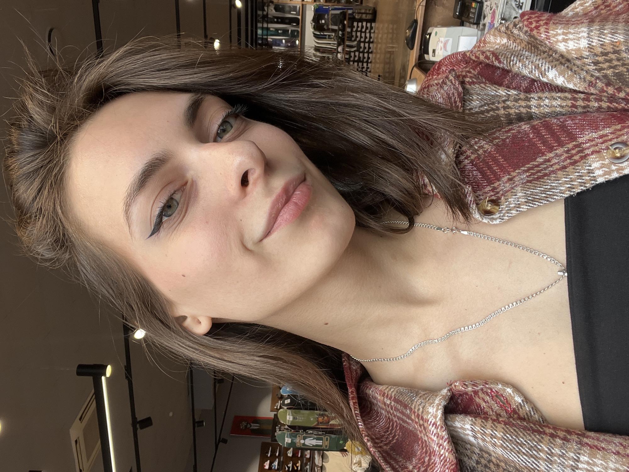 A young woman with fair skin and brown hair is the primary subject of the image. She is wearing a plaid shirt over a black top and a silver necklace. Her makeup includes eyeliner. The background appears to be an indoor setting. It has rows of items on shelves and overhead lighting. Skateboards are visible in the background. The location is Krasnoyarsk, Russia.