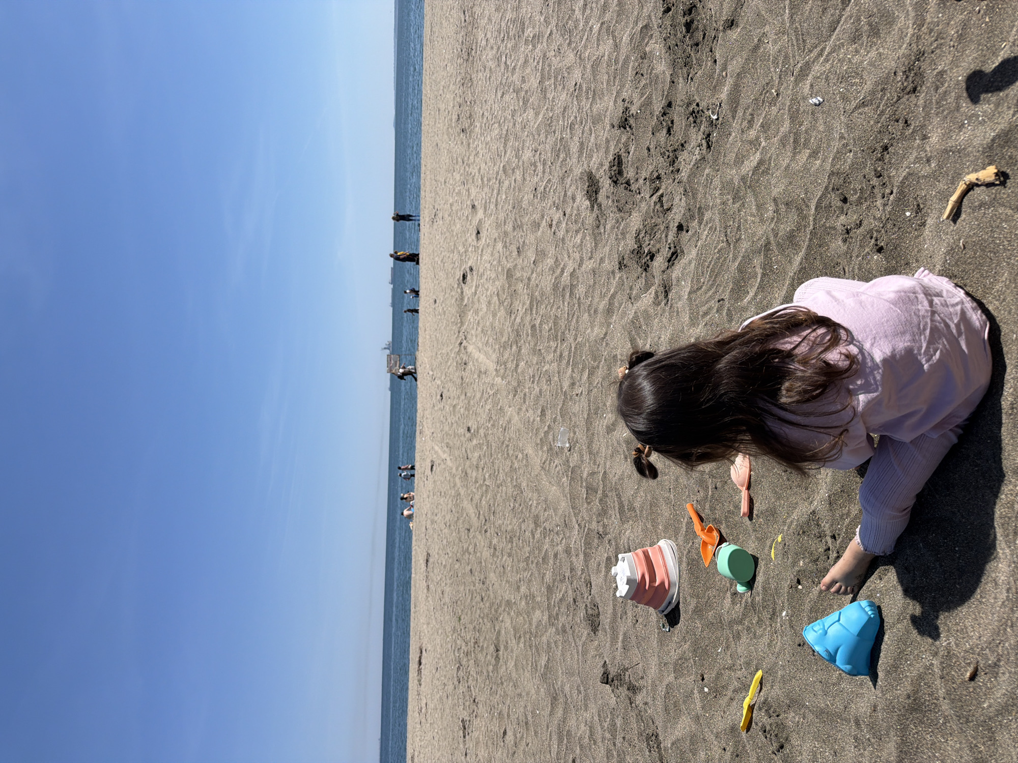 A young child with long, dark hair sits on a sandy beach on a sunny day in an Unknown city. The child is dressed in a light purple shirt and striped pants, and is surrounded by various beach toys including buckets, shovels, and molds. The beach stretches out towards the ocean, where several people are visible in the distance. The sky is clear and blue. The location is in the city of Unknown, Unknown.