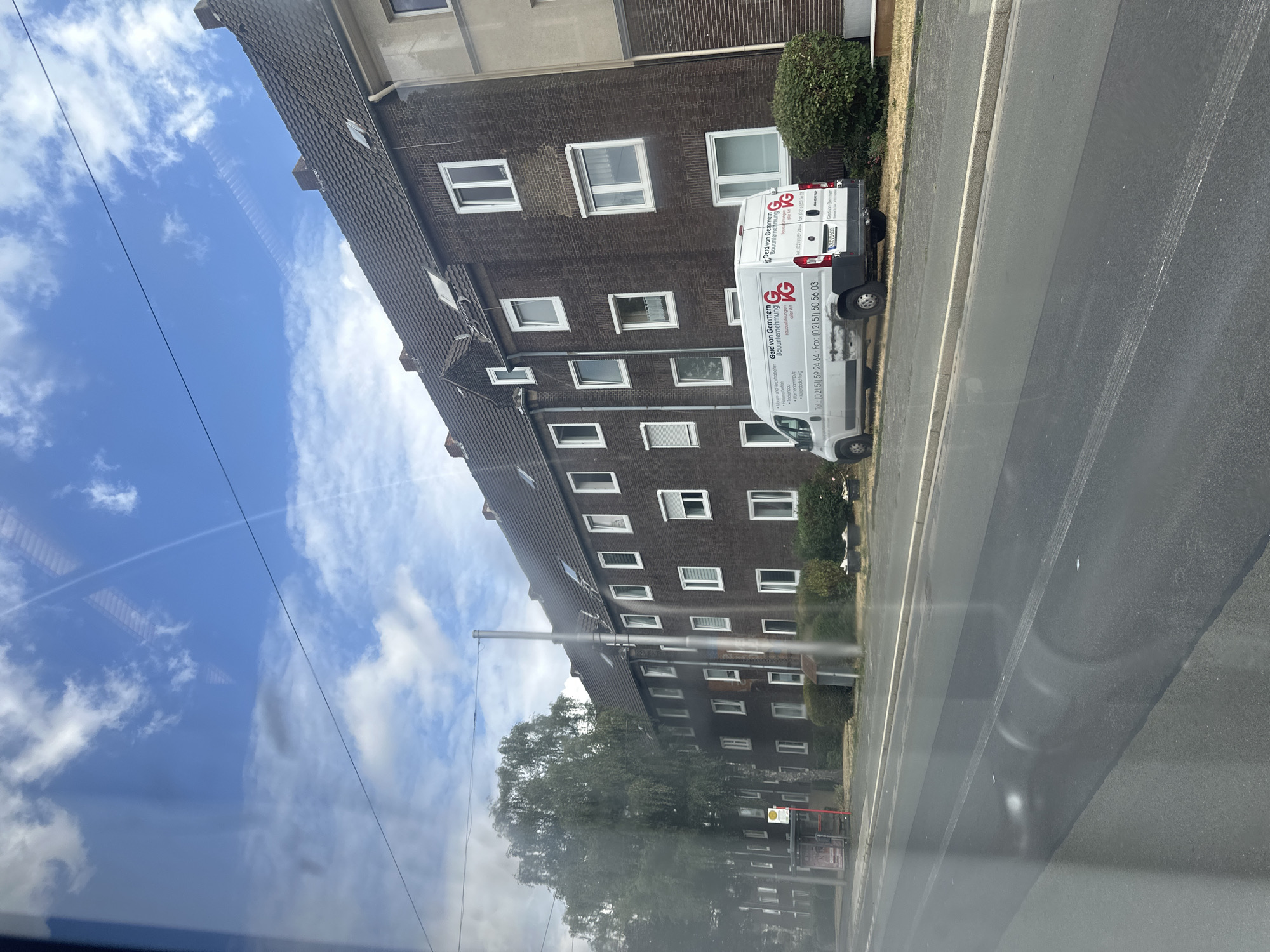 The image shows a street view in Krefeld, Germany, on a cloudy day. A multi-story brick apartment building with dark tiled roofs lines the right side of the street. The building has numerous windows, some with white frames and shutters. In front of the building, parked on the asphalt, is a white delivery van with the logo "VG" and company information printed on its side. To the left of the building, there is a bus stop shelter and a row of trees. The street in the foreground appears to have recently undergone some resurfacing or repair, with visible tire marks and some lighter patches on the asphalt. The sky above is blue with scattered white clouds.