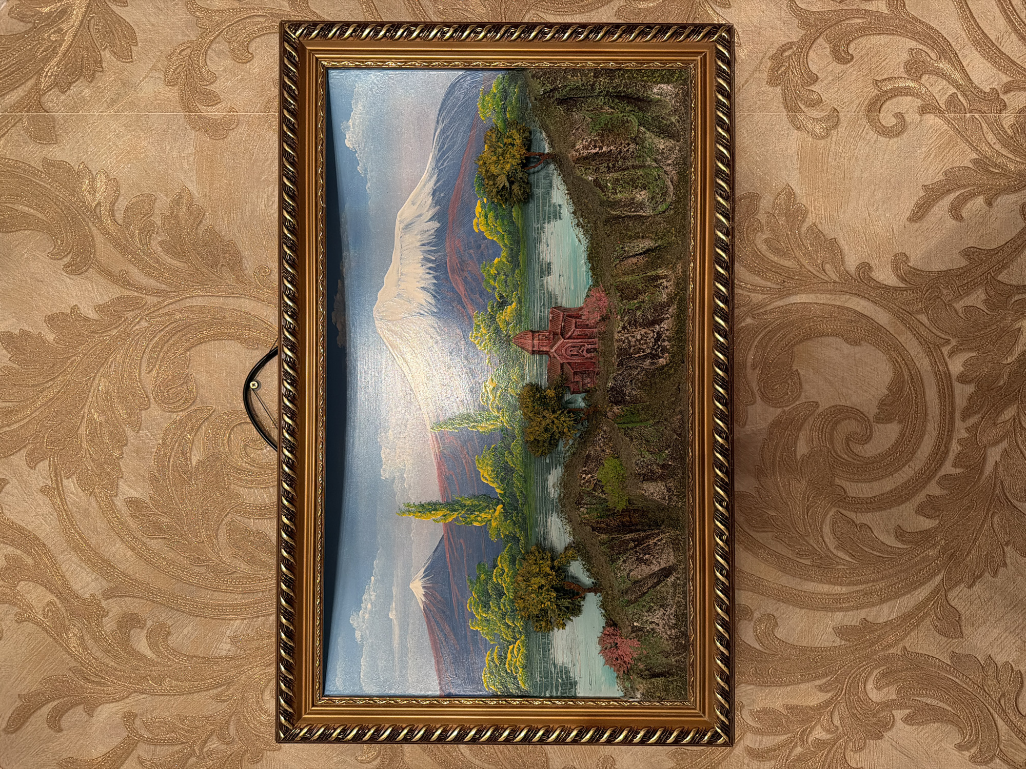 A framed painting hangs on a wall with patterned wallpaper. The painting depicts a landscape with a mountain in the background, trees, a body of water, and a building in the middle ground. The painting appears to have a textured or three-dimensional effect in certain areas, such as the trees and the building. The frame is ornate and gold-colored. The location is likely in Russia.