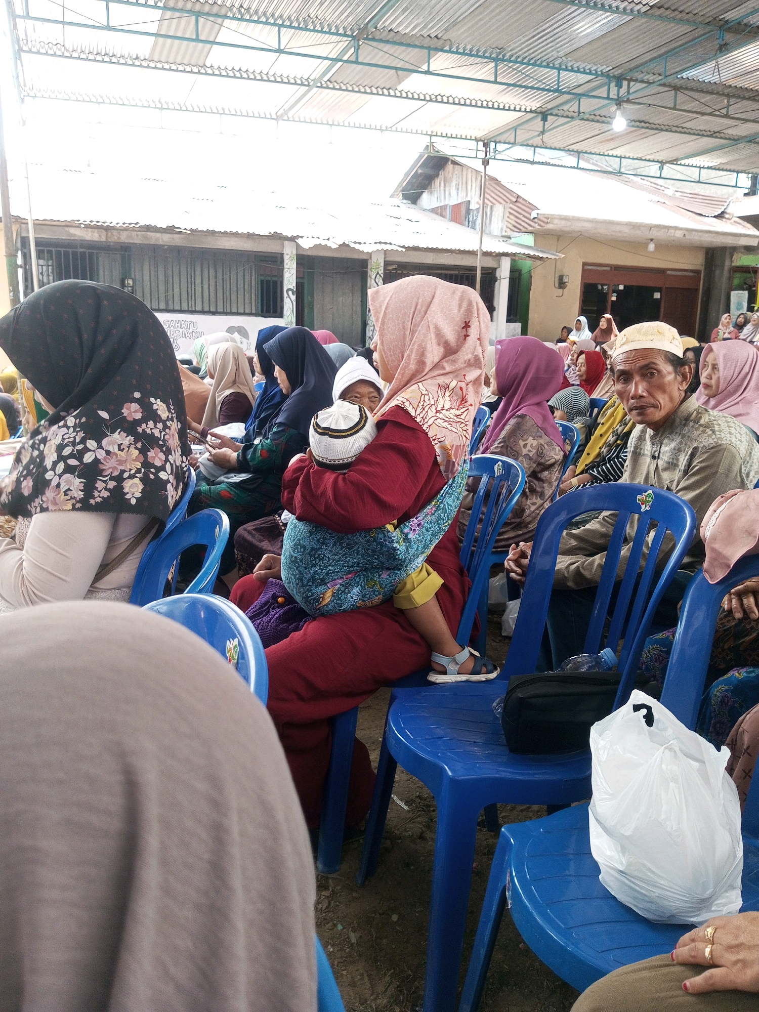 In Andongsili, Indonesia, under a corrugated tin roof supported by a metal frame structure, a group of people are seated on bright blue plastic chairs, facing a presumed stage or event area. The setting appears to be outdoors or semi-outdoors, with a rough dirt floor. The lighting is bright, suggesting daytime.
In the foreground, a person wearing a dark hijab with a floral pattern is seated. To their right, a woman in a pink hijab is holding a baby wrapped in a blue and patterned cloth, with the baby wearing a white and black prayer cap. The woman is also wearing a red garment.
Further back and to the right, a man in a light-colored patterned shirt and a white cap is looking directly at the camera, his hands clasped. He is seated on a blue chair. Adjacent to him, another person in a pink hijab is visible.
Several other individuals are seated in the rows of blue chairs, mostly wearing hijabs and modest clothing, indicating a cultural or religious gathering. Objects on the chairs include a black bag and a white plastic bag, likely containing belongings. The overall scene depicts a community event or gathering in a somewhat rural or modest setting.