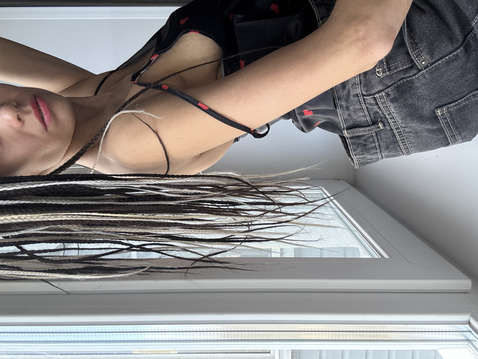 A woman is lying horizontally near a window. She has long braids in varying shades of brown and blonde. She's wearing a black top with red heart patterns and dark denim jeans. The background features a white window frame. The image does not provide sufficient information to definitively confirm the location in the city of Rusanovo, Russia.
