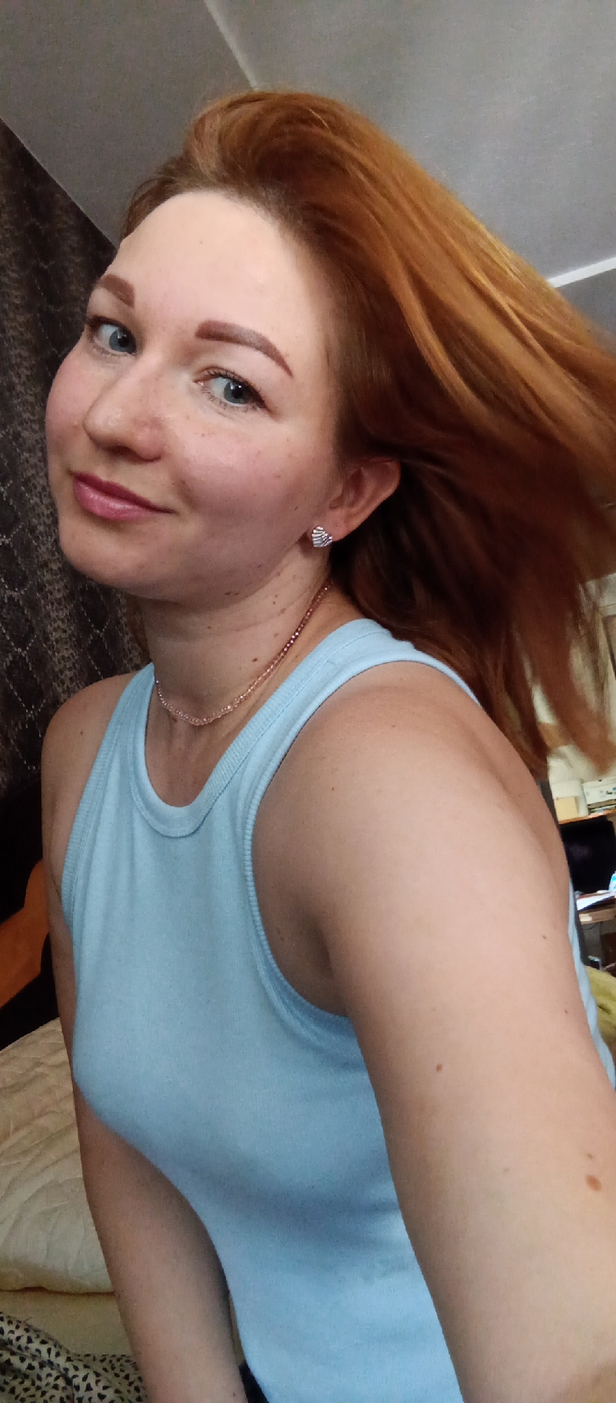 A selfie taken indoors depicts a woman with red hair and blue eyes. She wears a light blue tank top and a delicate pink necklace. Her hair is in motion, swept to the side. She has fair skin with freckles and is wearing stud earrings. The background features a patterned wall and a glimpse of a bed with light-colored bedding. Part of a desk with a computer or monitor can also be seen. The location is Unknown, Unknown.