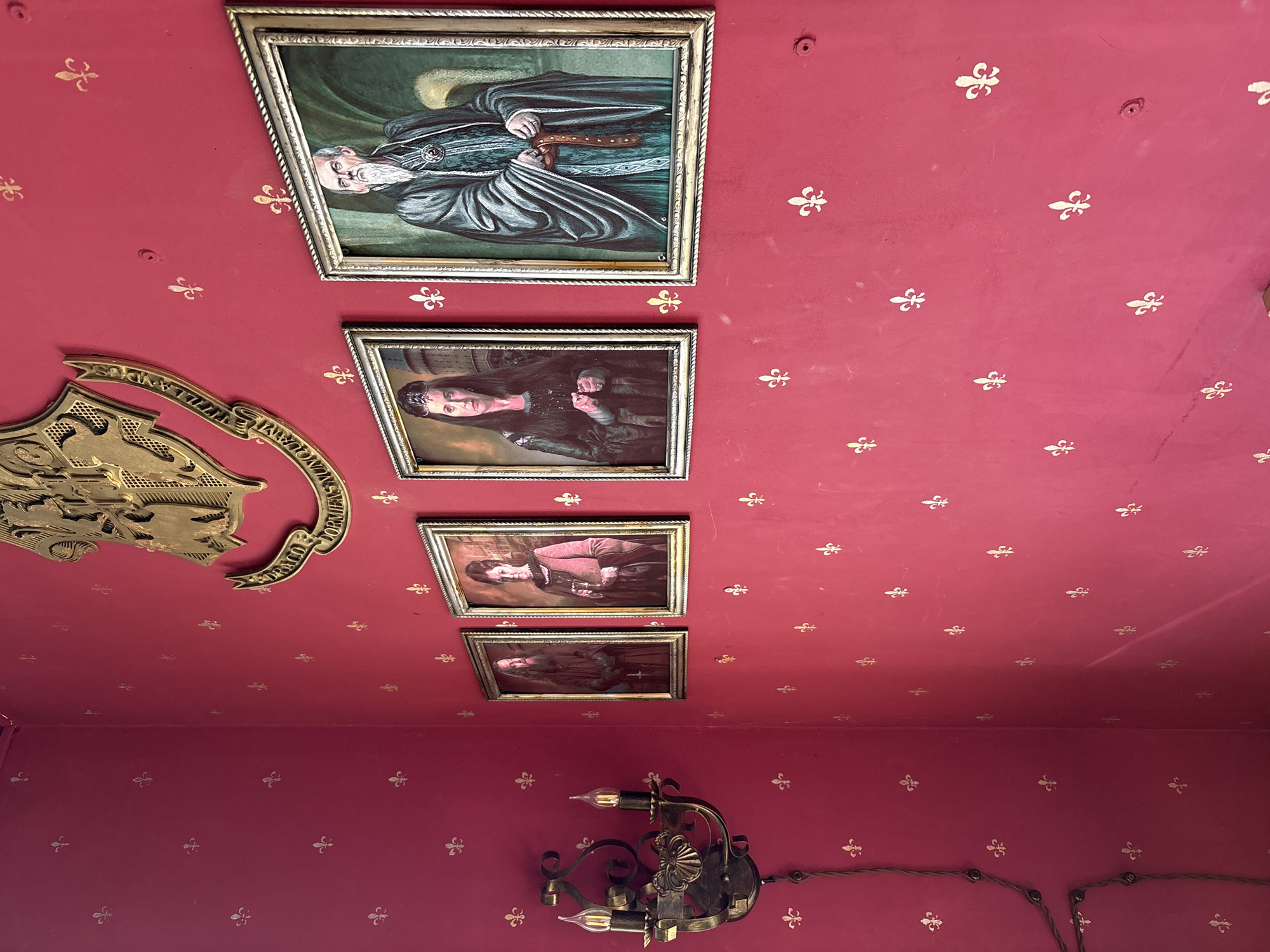 The image shows a room in Kazan, Russia, with a red ceiling and wall adorned with a gold fleur-de-lis pattern. Framed portraits hang on the ceiling. From top to bottom: a man with a beard in dark robes; a woman in a dark dress; a woman in a pink dress; and a person in dark clothing. A gold shield-shaped emblem is mounted on the upper left of the ceiling. An ornate metal wall sconce with two exposed light bulbs is attached to the bottom of the wall on the left side. The ceiling and wall appear to meet at a 90 degree angle. A long electrical wire is stretched across the wall.