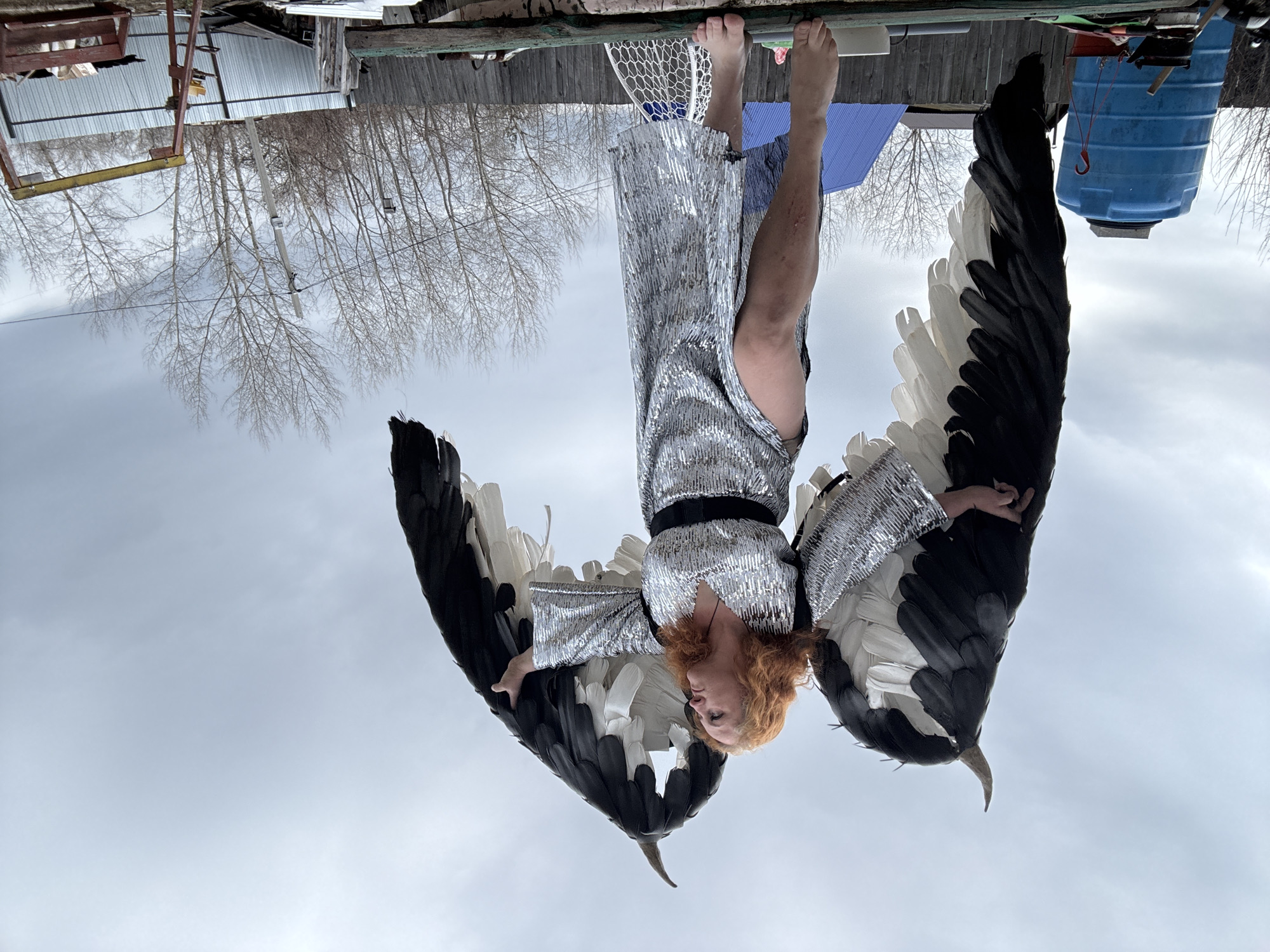 A woman is suspended upside down wearing a silver sequined dress, a black belt, and large black and white wings. She has red hair and fair skin. Her feet are visible above her head. The background consists of a drab, overcast sky, bare trees, and dilapidated structures including a corrugated metal building, a blue plastic water tank, and other unidentified objects. The scene may be in the city of Chaikovsky, Russia.