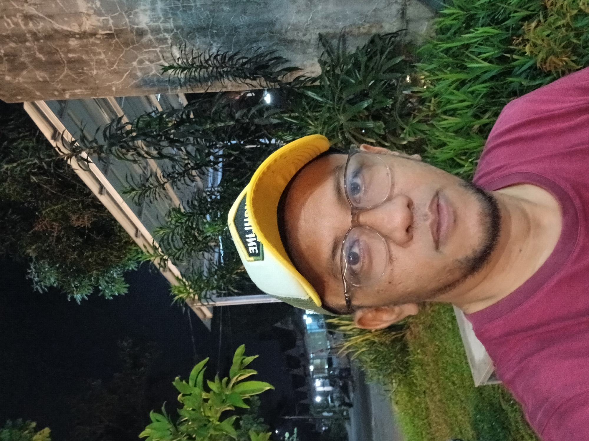 The image is a close-up, selfie-style photograph of a man wearing a maroon t-shirt and a yellow and white baseball cap. He is also wearing clear-rimmed glasses and has a light beard. He is outdoors at night, with trees, foliage, and a textured wall visible in the background. In the distance, there are lights from buildings and parked vehicles. The overall setting appears to be a public space, possibly a park or a street in a city, with residential buildings suggested in the background. The image was taken in Kebalen, Indonesia.