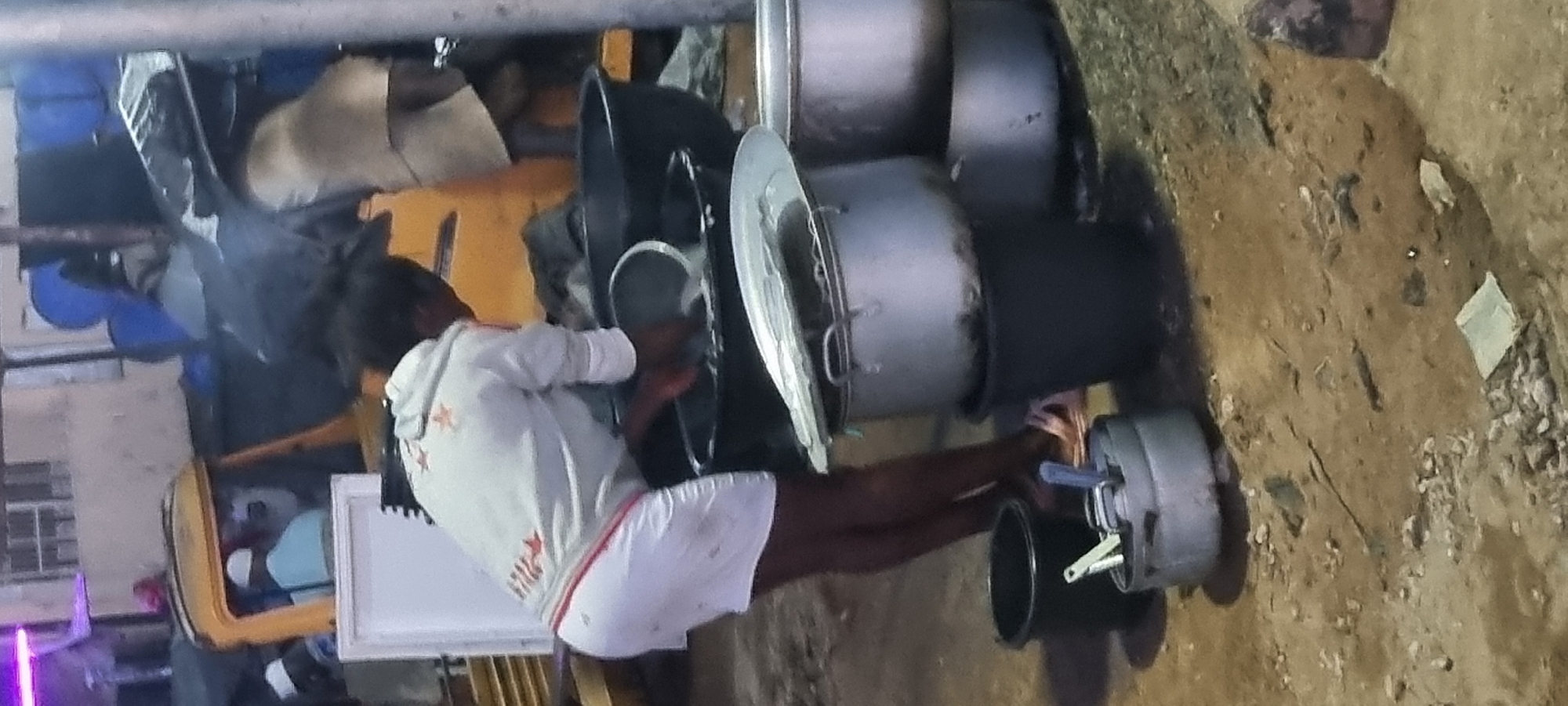 A person in a white hoodie and shorts stands near a collection of pots and cooking equipment outdoors. The person is interacting with the cooking utensils. The location appears to be a street scene. Based on the lighting, it is likely nighttime. This is a roadside food vending location in Lagos, Nigeria.