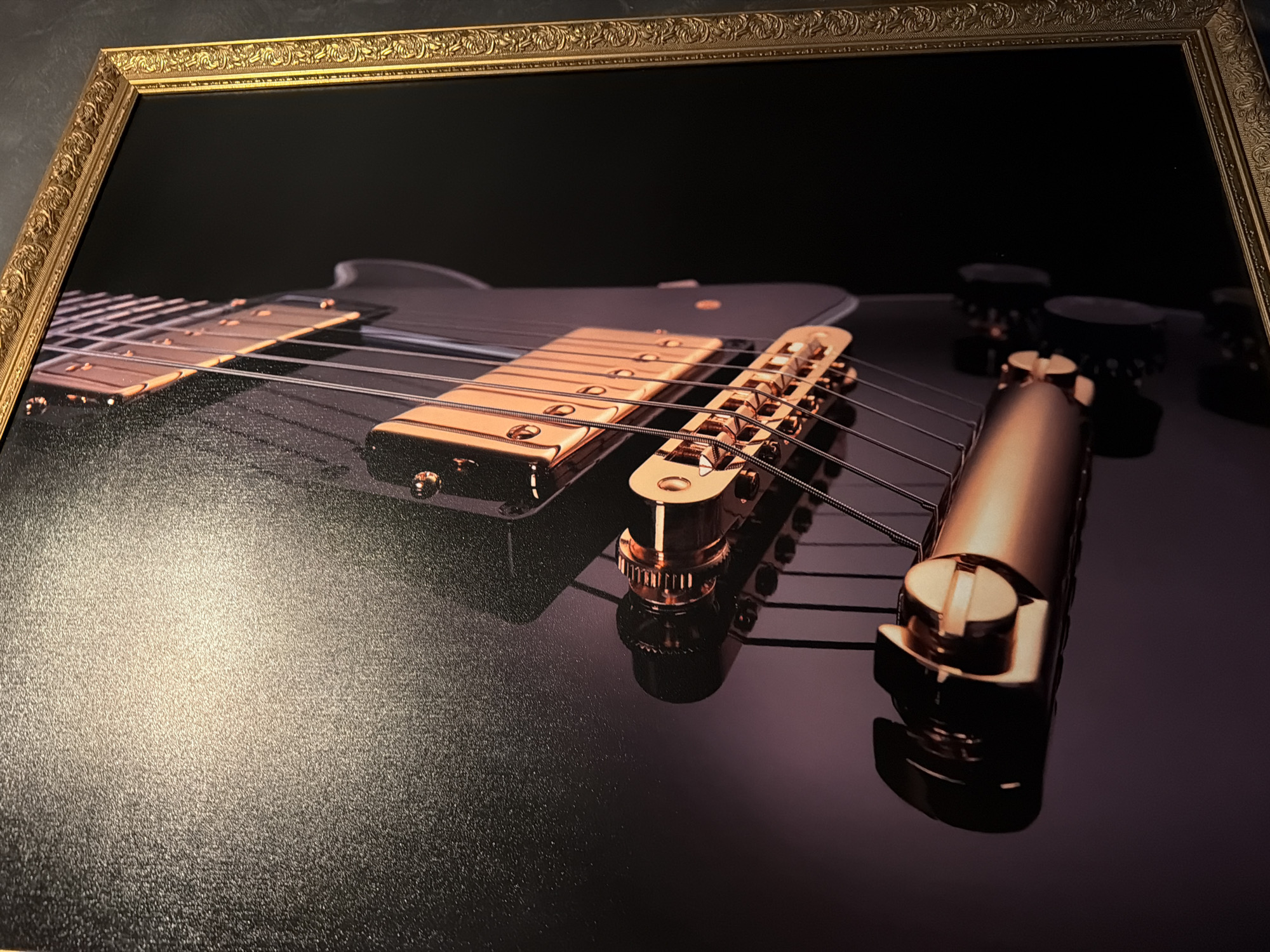 A framed photograph depicts a close-up view of an electric guitar, focused on the strings, bridge, and knobs, set against a black background. The frame is ornate and gold. The metallic components of the guitar are a polished gold color, reflecting light. The strings are taut and traverse from the bridge toward the headstock. The knobs, visible in the background, appear to be black. The exact location is Unknown, Unknown.