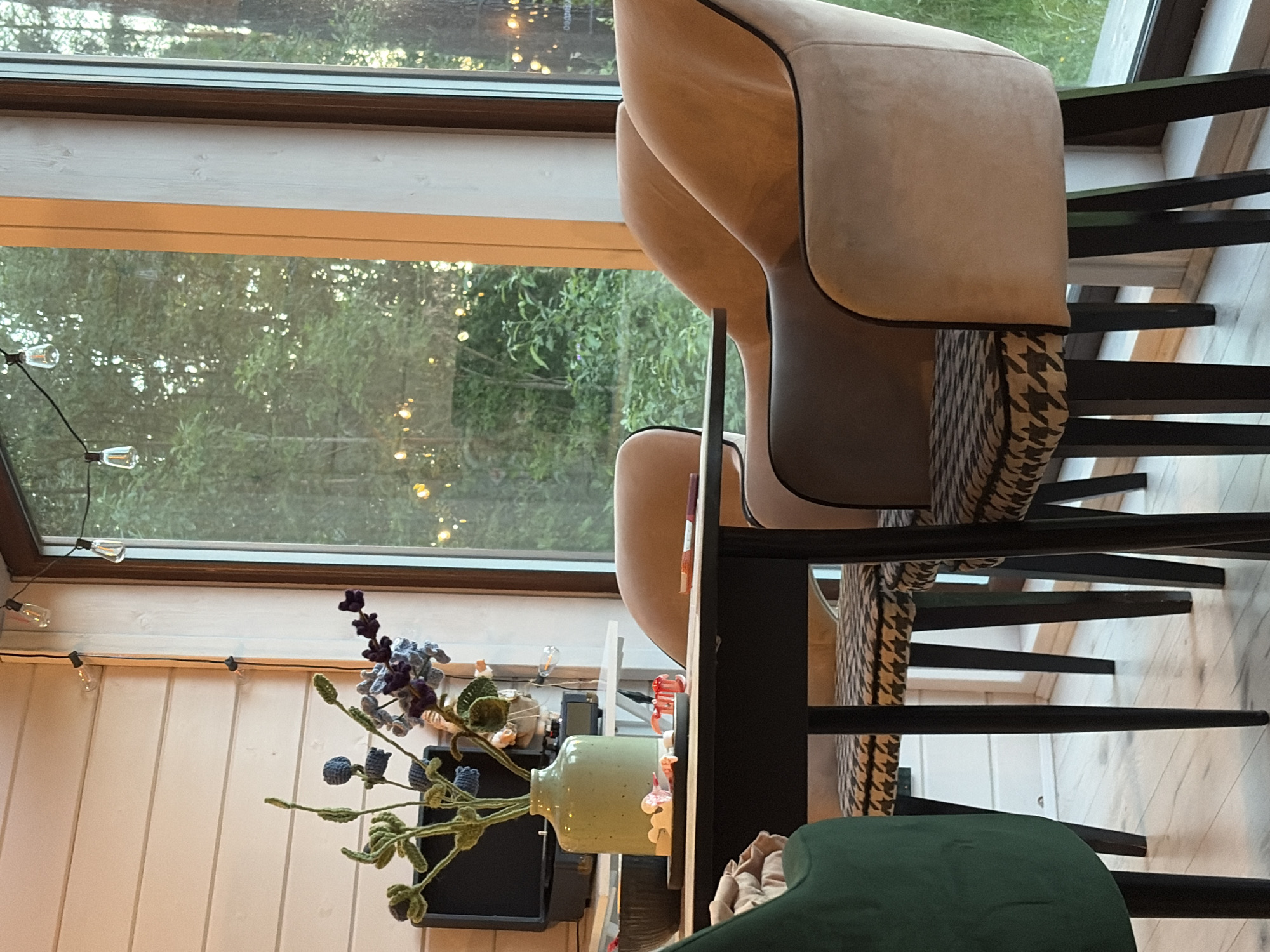 The image depicts a dining area with a table and chairs positioned in front of a large window. The window overlooks a natural, tree-filled landscape, with the soft glow of string lights visible in the distance, suggesting an outdoor evening setting. Inside, the room features light-colored wooden paneling on the walls and a light-colored wooden floor.
On the table, there is a light green ceramic vase containing a bouquet of artificial flowers. Beside the vase, a black rectangular tray holds several small, light-colored objects that appear to be candies or decorative items, along with a small red decorative item. A dark-colored bowl is partially visible on the left side of the table.
There are three chairs visible around the table. One is a dark green upholstered chair on the far left, mostly out of frame. Two other chairs are positioned to the right, with dark wooden legs. These chairs have light beige upholstered backs and sides, and seat cushions with a black and white houndstooth pattern. The arrangement suggests a dining setting for at least three people.
In the background, on a white shelf, a black electronic device, possibly a record player or speaker, and a small figurine are visible. String lights with exposed bulbs hang from the ceiling along the wall. The overall atmosphere is cozy and inviting, with natural light filtering through the window and the warm glow of artificial lights. The specific location in Snegiryovka, Russia, is not evident from the image itself, but the setting evokes a sense of a rural or semi-rural retreat.