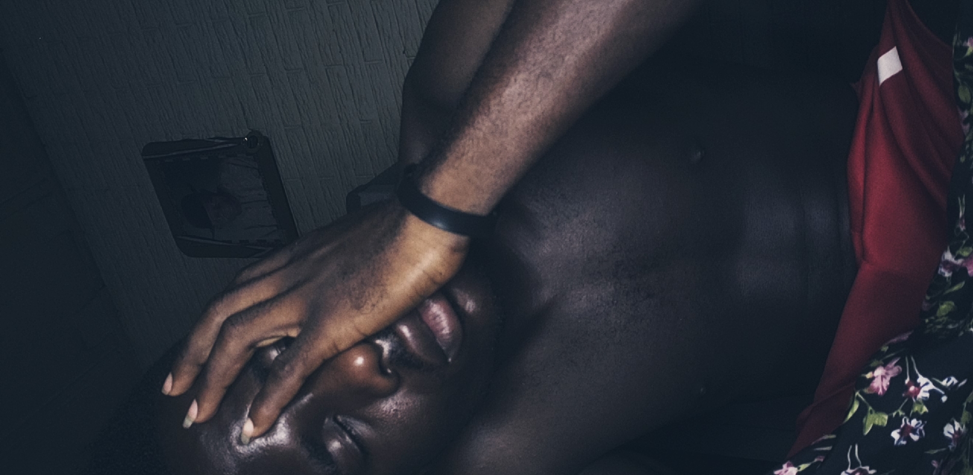 A dark-skinned individual is lying down in what appears to be a dimly lit room, possibly indoors. The person's face is partially obscured by their own hand, which is adorned with a black wristband. To the left of the frame, a rectangular object, perhaps a bag, is visible against a wall with a textured pattern. To the right, hanging garments in shades of red and black are present. There is no definitive information to confirm the location in Narayi, Nigeria.