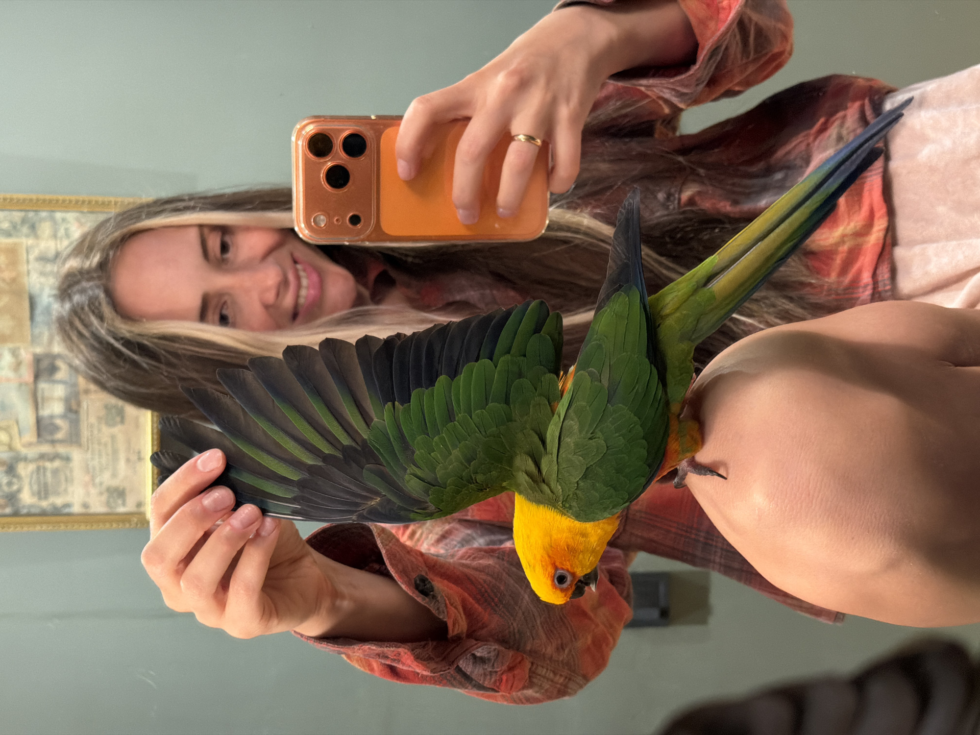 A woman is taking a selfie in a mirror with a sun conure parrot perched on her shoulder. The parrot's wings are spread, revealing green and black feathers. The woman is smiling and holding an orange-cased smartphone. She is wearing a red and orange plaid shirt. The background is a light blue wall with a framed picture hanging on it. The scene appears to be indoors, possibly in a bathroom or bedroom. There is no indication of the location being in Khimki, Russia.