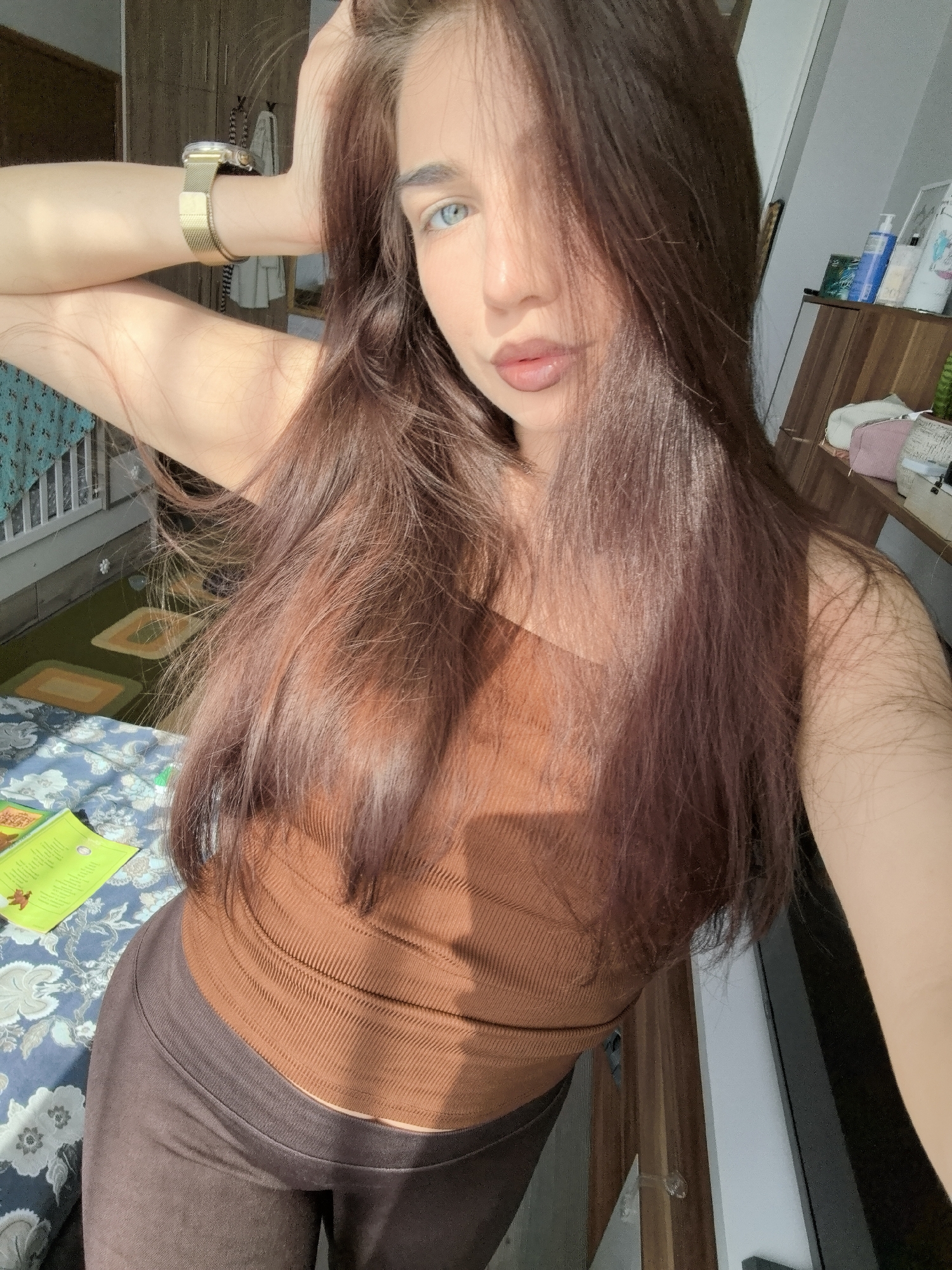 The image depicts a young woman taking a selfie indoors, likely in a bedroom or personal space, under bright natural sunlight in Stara Zagora, Bulgaria.
The main subject is a woman with long, dark reddish-brown hair, which partially covers her face. Her left eye, a striking light blue, is clearly visible and looks directly at the camera. She has full, neutral-toned lips. She is dressed in a fitted, ribbed, sleeveless brown top and dark brown leggings or pants. A gold-toned watch with a mesh strap adorns her left wrist, which is raised with her hand casually placed in her hair, creating a natural pose.
The setting appears to be a sunlit room. To the left, a bed is visible with a blue and white floral patterned duvet or bedspread, and a green book or notebook rests on it. Behind the bed, a light-colored railing and a patterned curtain can be seen, along with a green rug featuring geometric shapes on the floor. To the right, a wooden cabinet or dresser holds various personal items, including several bottles (one blue bottle visibly labeled "SEBORADIN SHAMPOO" and "CONDITIONER"), a small plant, a pink pouch or purse, and a white-framed item. Strong sunlight streams into the room from the left, casting shadows and highlighting parts of her hair and arm, indicating it is daytime. The overall impression is casual and relaxed.